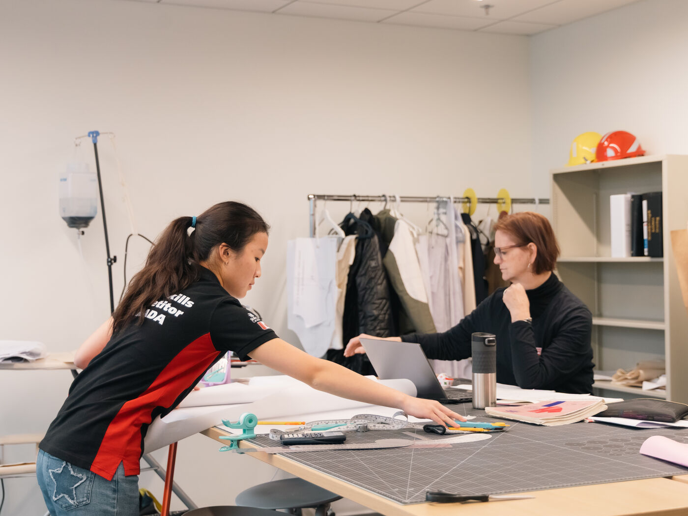 Two women are working on a project, one using a laptop and the other pointing at something on a desk with design materials.