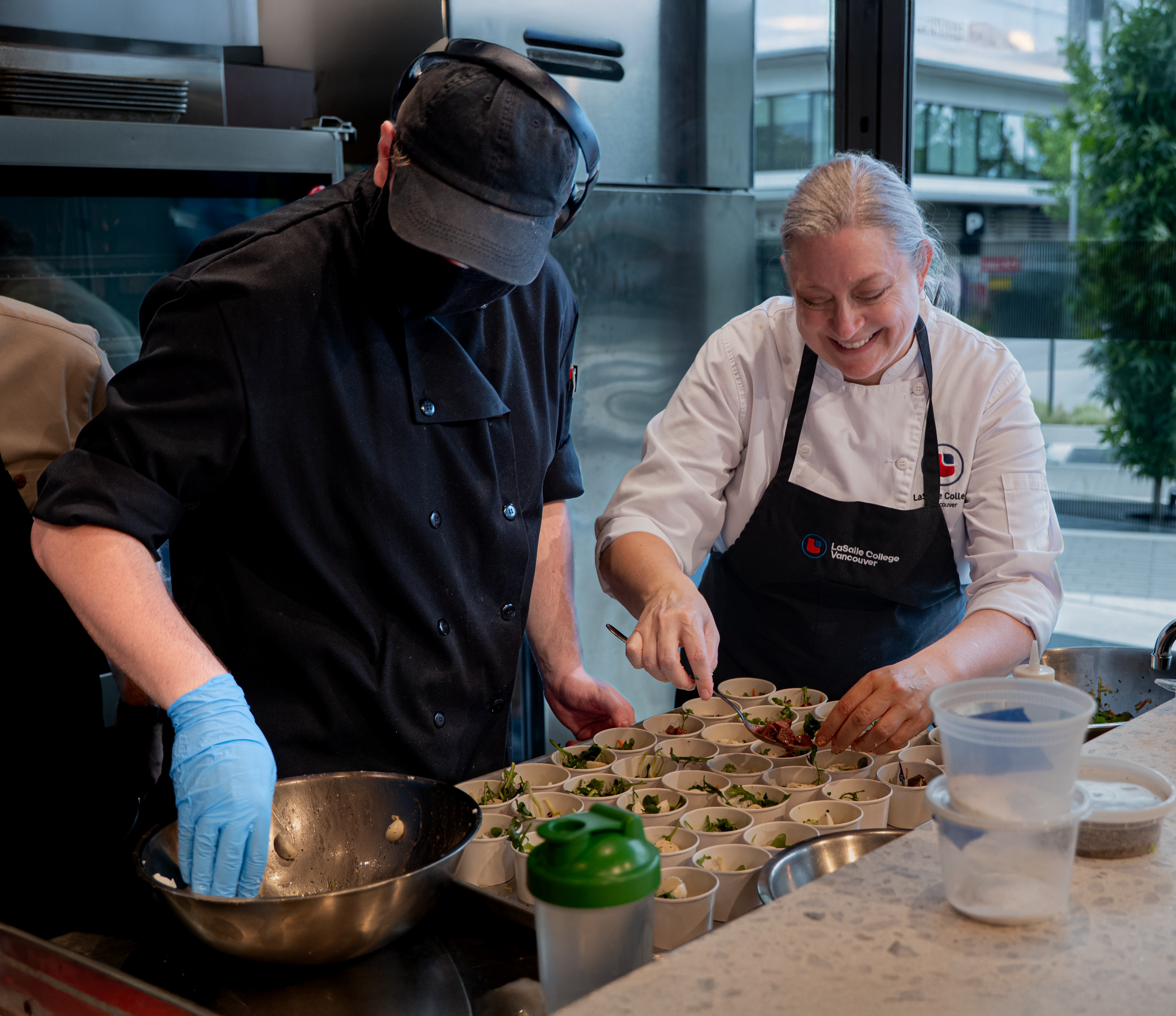 Two chefs are preparing food in a professional kitchen setting, carefully placing ingredients into small containers for an event or service.