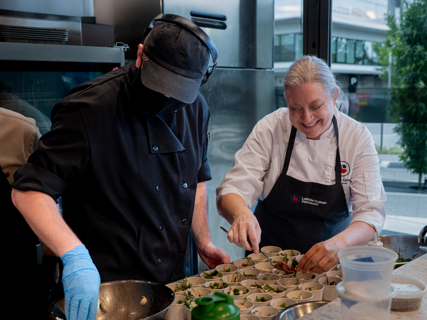 Two chefs are preparing food in a professional kitchen setting, carefully placing ingredients into small containers for an event or service.
