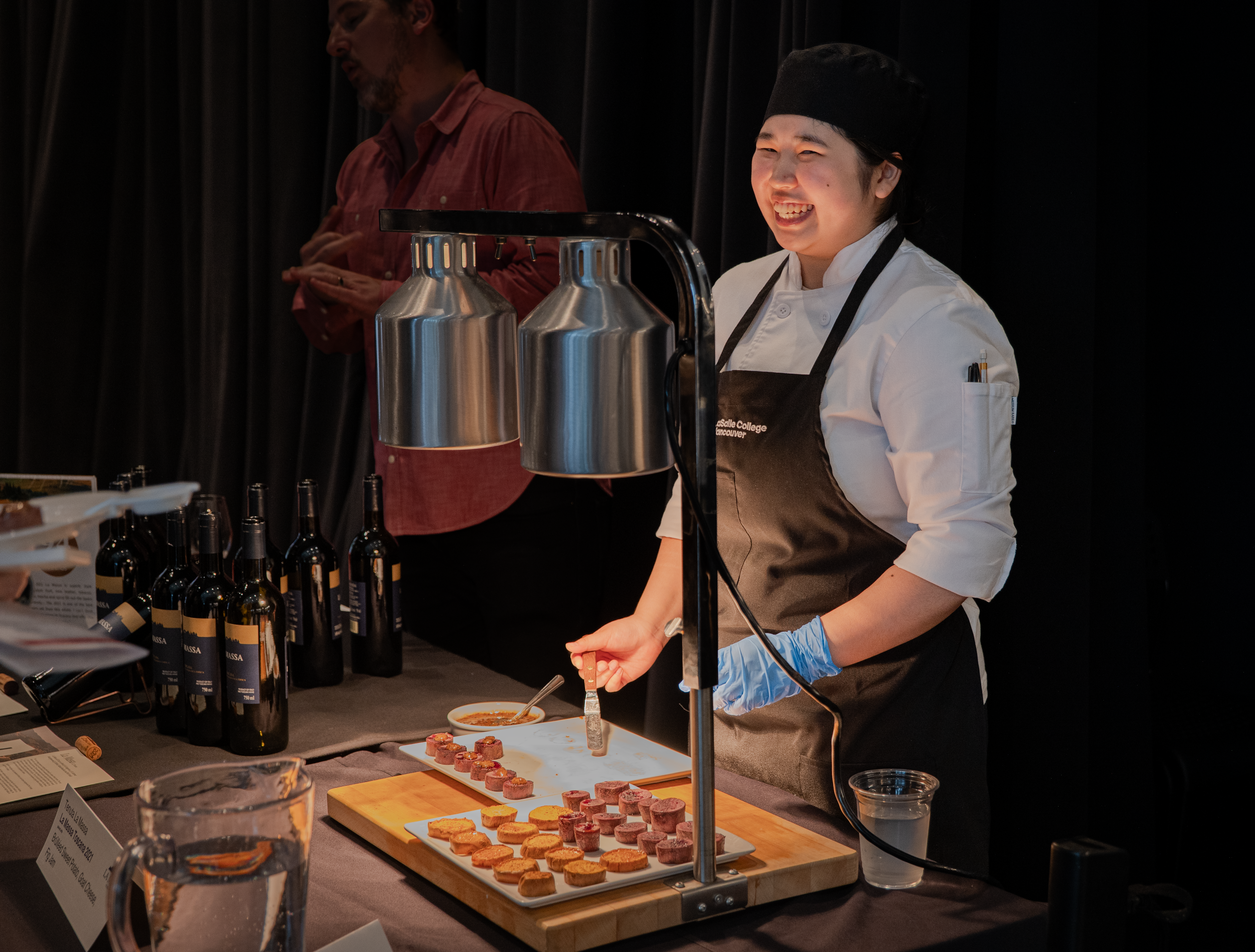 A smiling chef serves appetizers at an event, with wine bottles and other preparations on the counter.