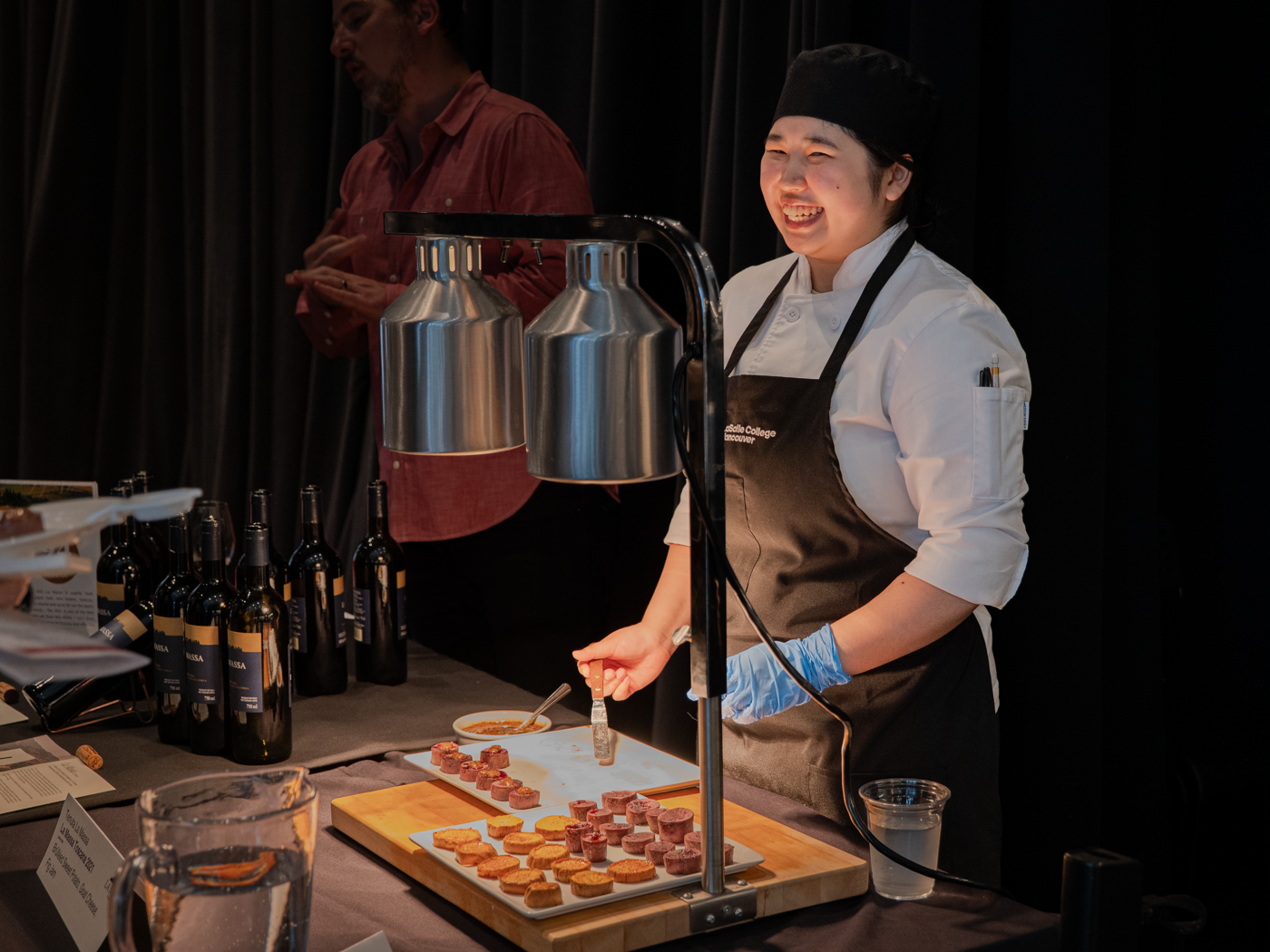 A smiling chef serves appetizers at an event, with wine bottles and other preparations on the counter.
