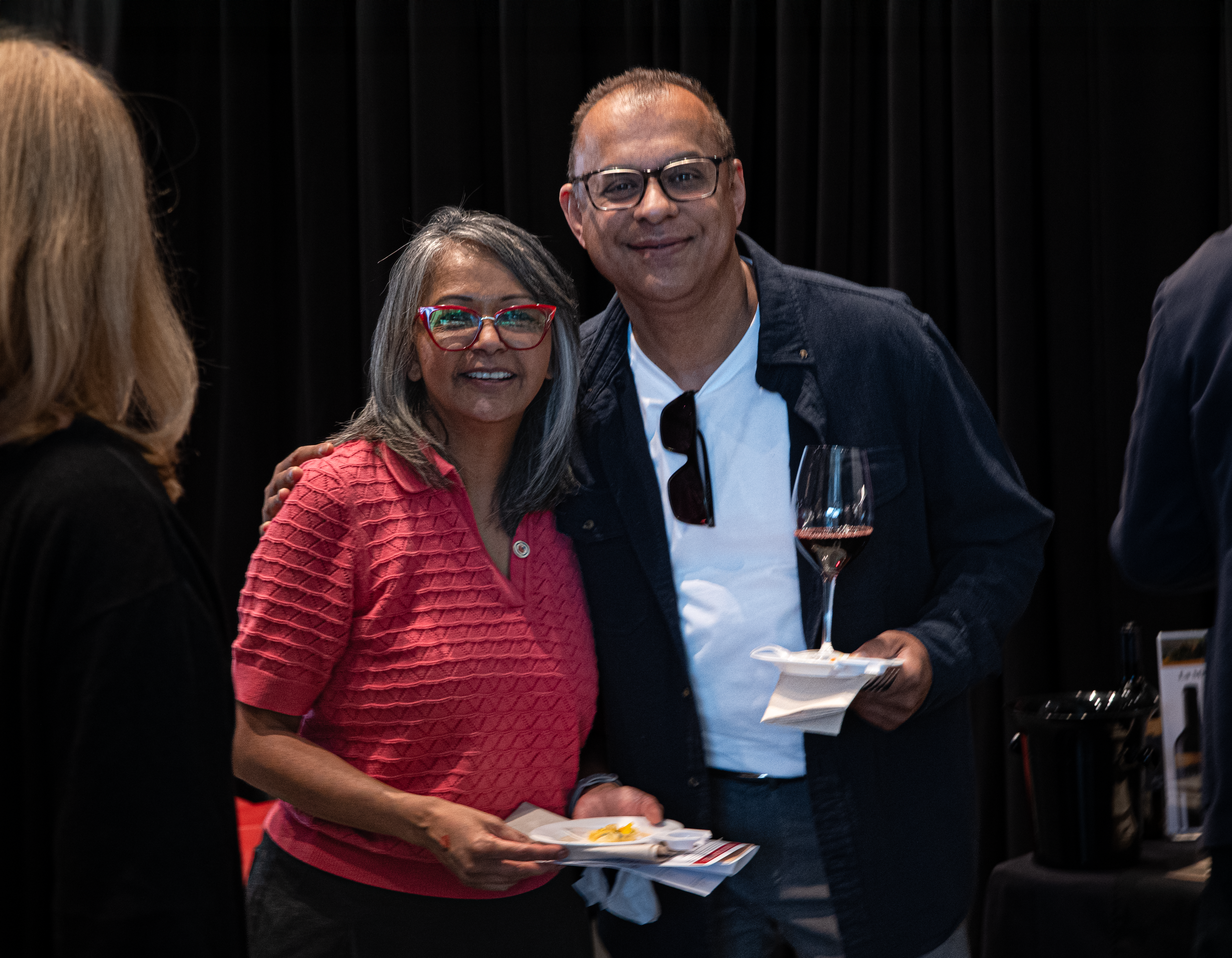 A woman and a man smile at the camera, she is wearing a red blouse and he is wearing a dark jacket holding a glass of wine.