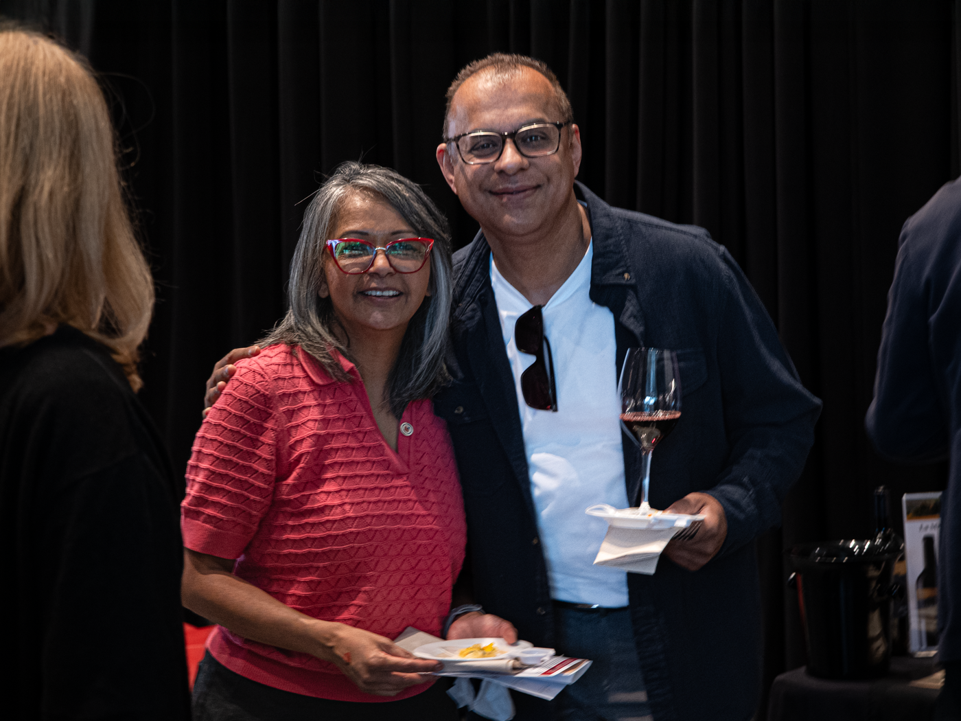A woman and a man smile at the camera, she is wearing a red blouse and he is wearing a dark jacket holding a glass of wine.