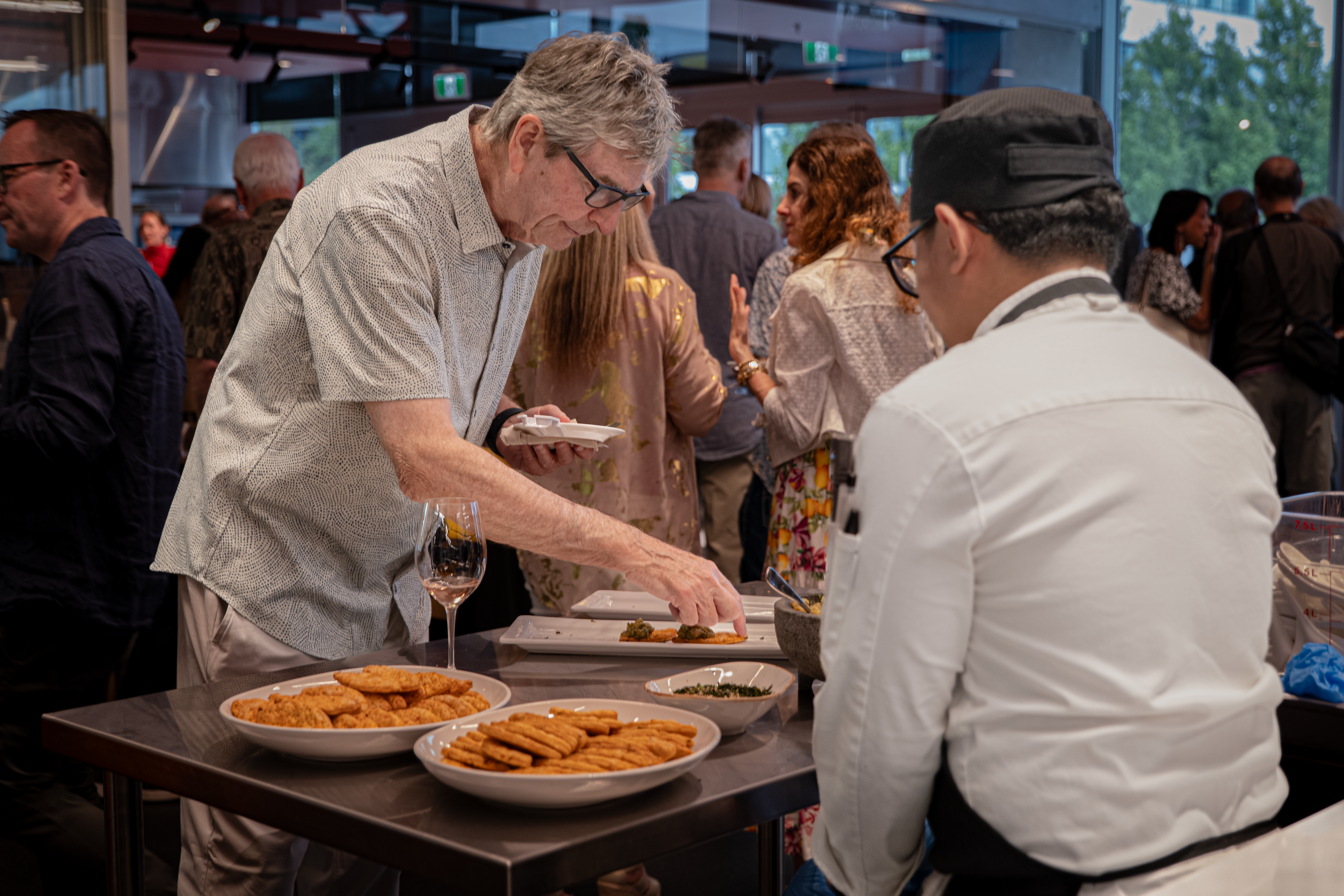 An older man serves food from a platter to another person while other attendees at a social event enjoy the food and company.