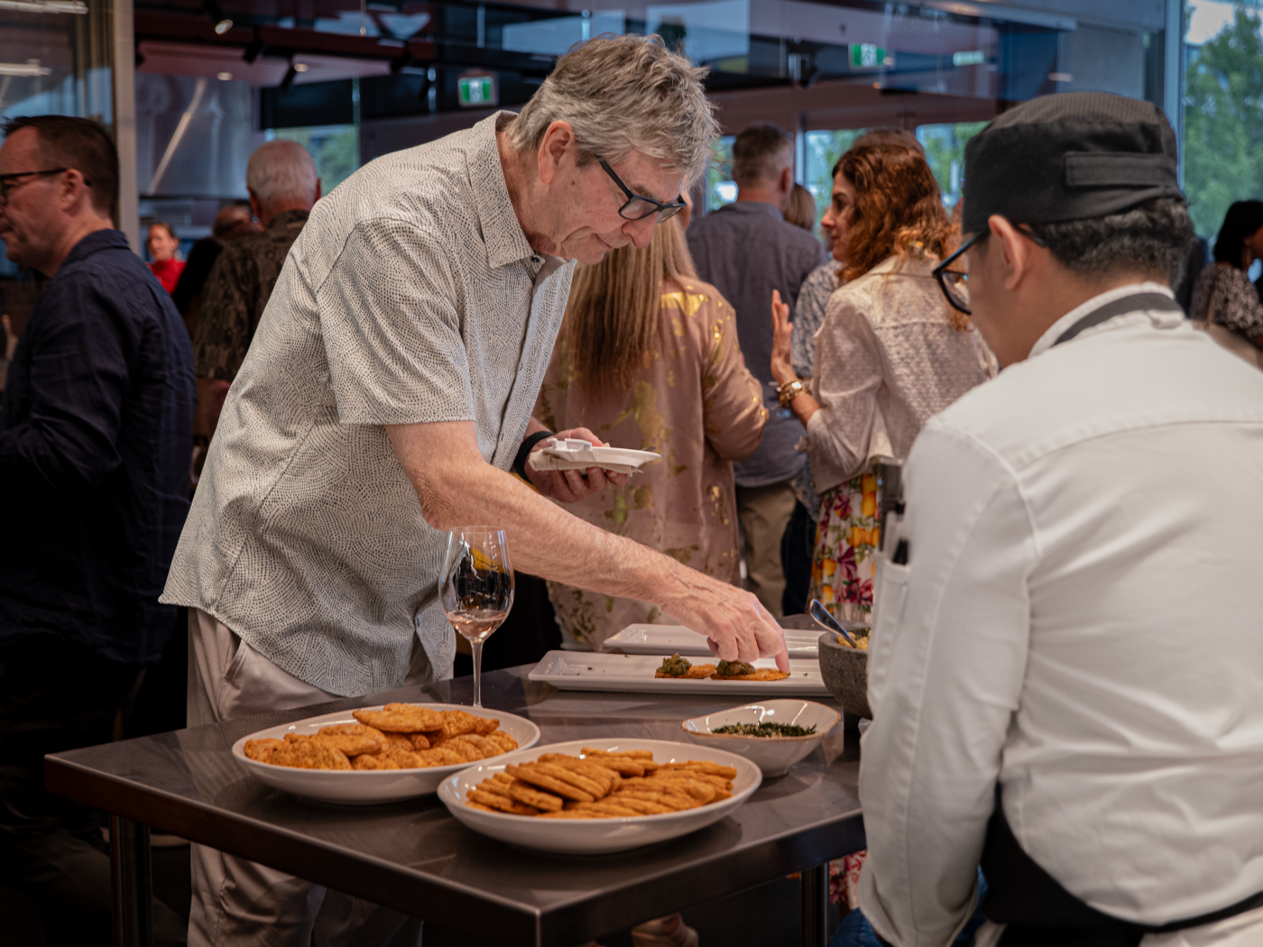 An older man serves food from a platter to another person while other attendees at a social event enjoy the food and company.