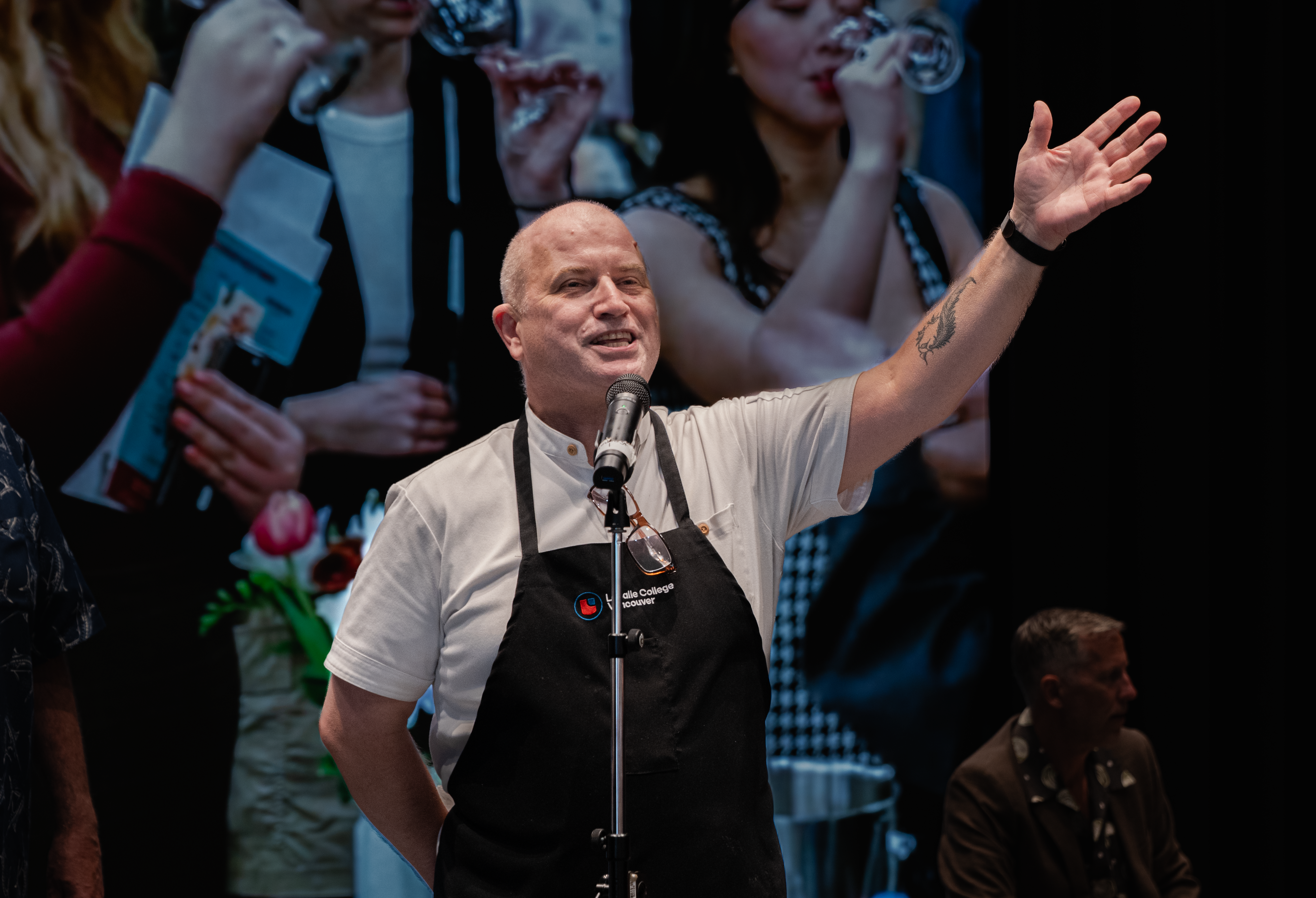 A bald man wearing a black apron speaks into a microphone, with one hand raised in a gesture. In the background, women are seen drinking wine.