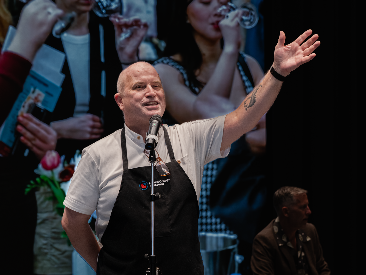 A bald man wearing a black apron speaks into a microphone, with one hand raised in a gesture. In the background, women are seen drinking wine.