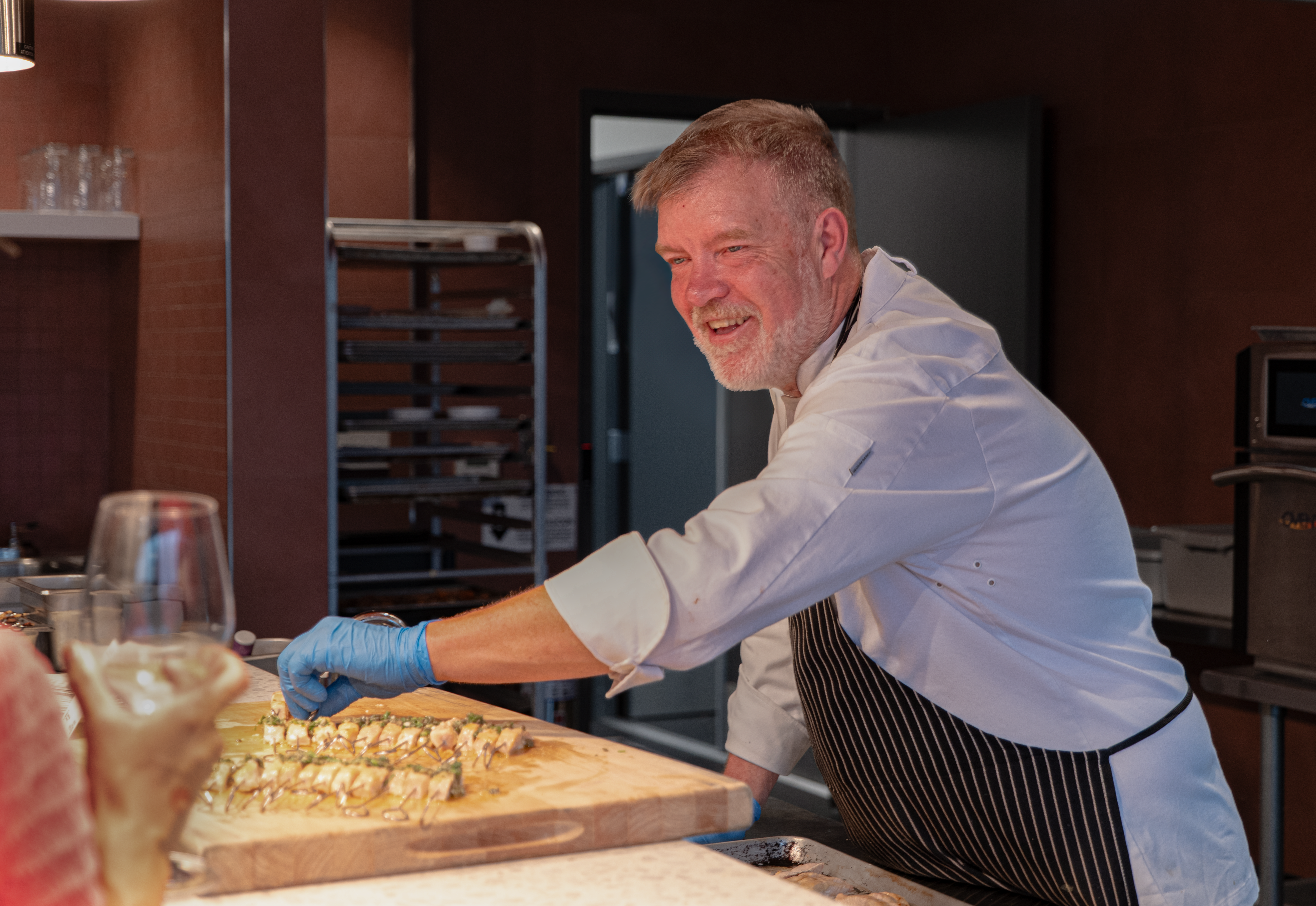 A smiling chef wearing an apron and chef's hat is preparing sushi rolls with blue gloves on in a professional kitchen.