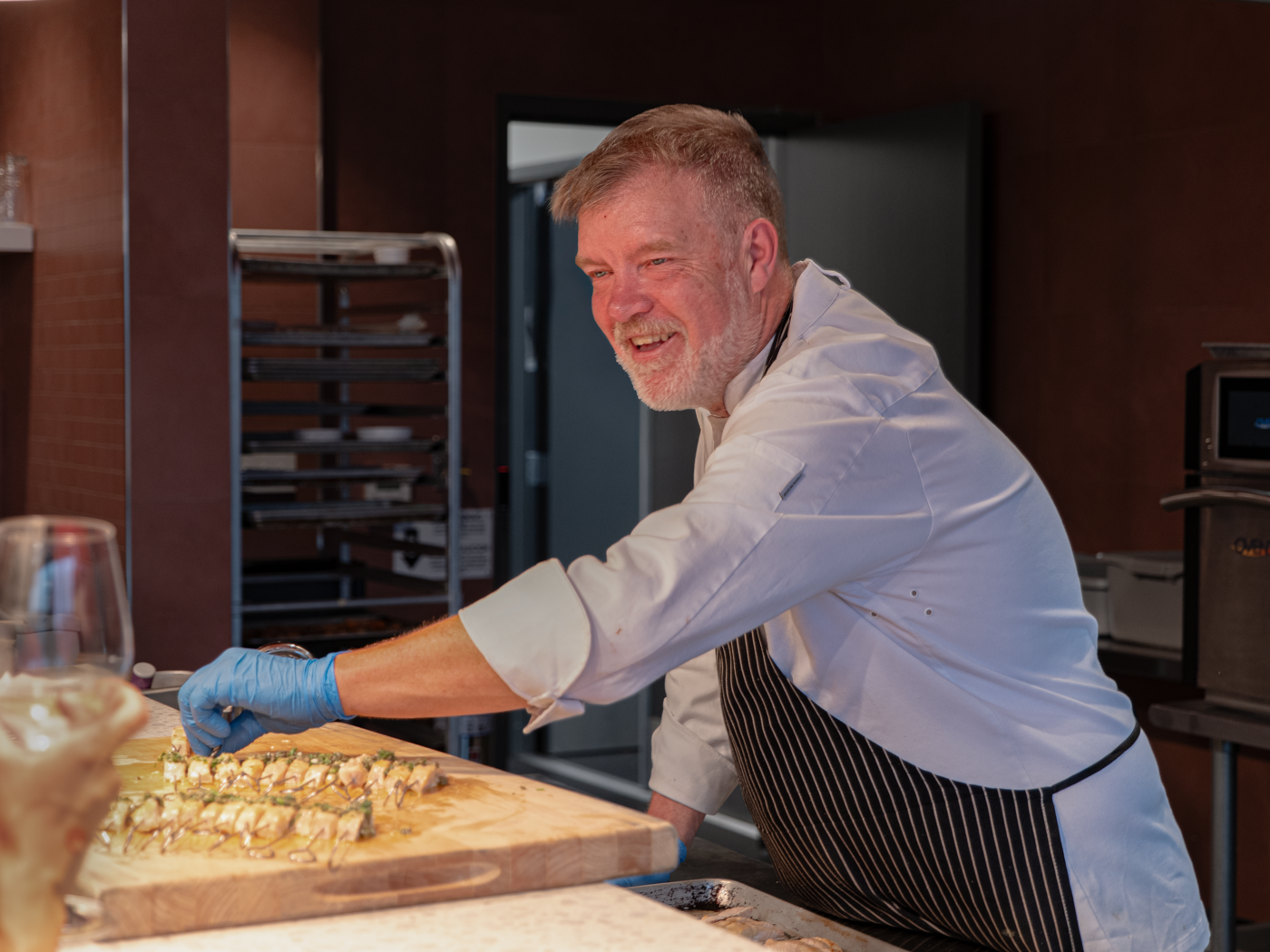 A smiling chef wearing an apron and chef's hat is preparing sushi rolls with blue gloves on in a professional kitchen.