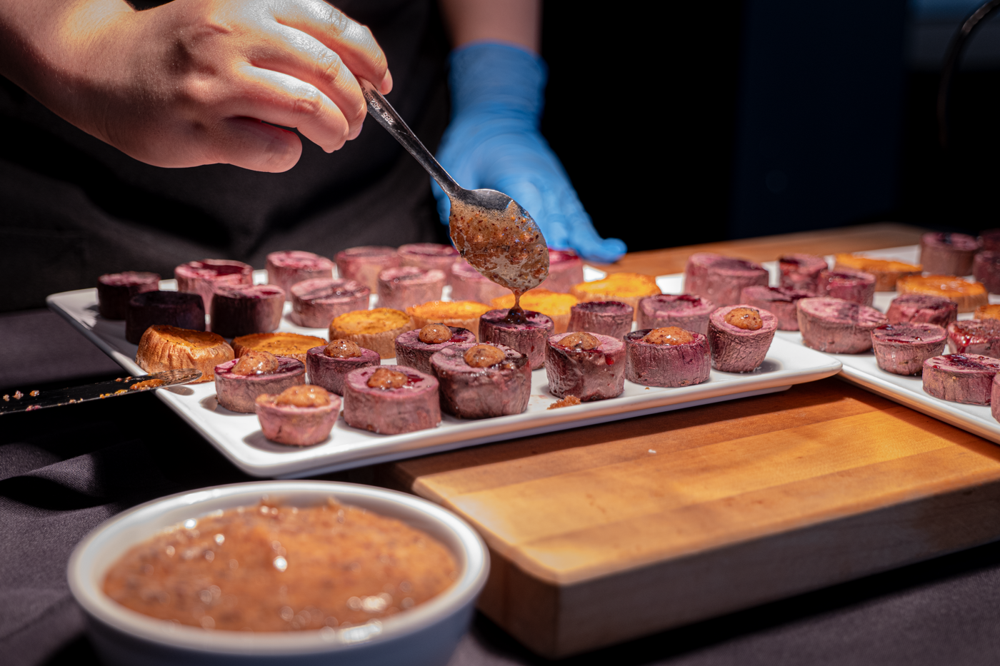 A person wearing blue gloves is decorating small purple pastries with a spoon of sauce, presented on white platters.