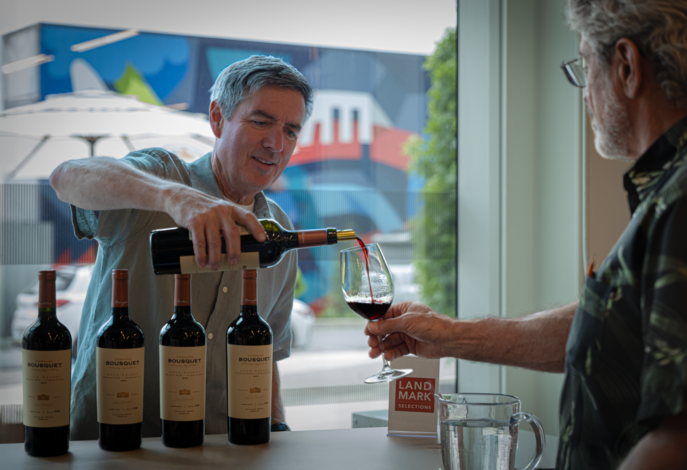 A man pours red wine from a bottle into a glass as another man watches. There are four bottles of wine on the table.