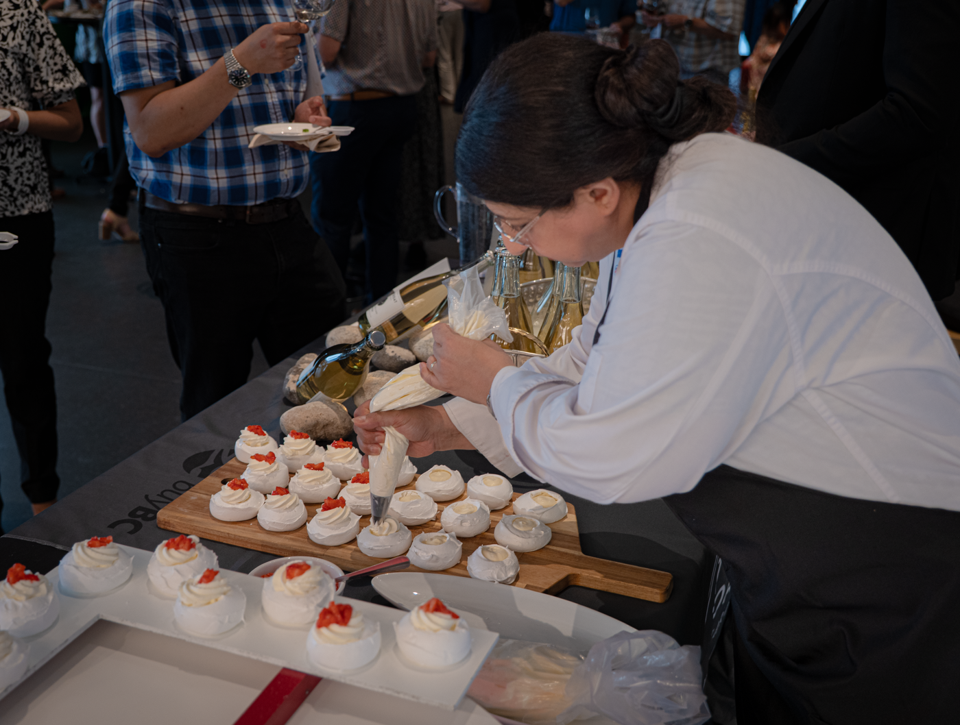 A female chef is meticulously decorating meringue desserts with red fruit using a piping bag, with wine bottles and glasses visible in the background.