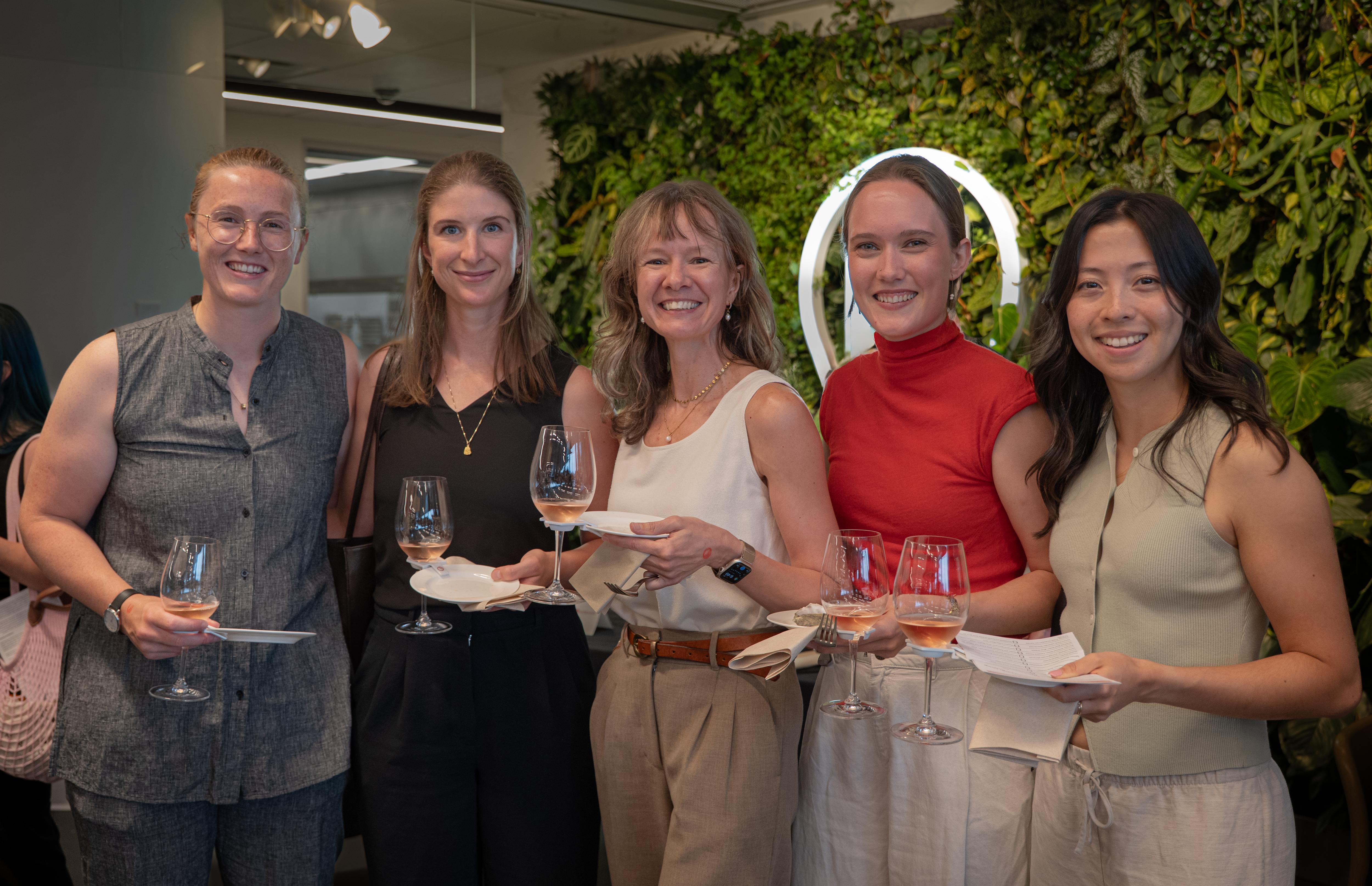 Five smiling women holding wine glasses and appetizers pose in front of a green wall with an illuminated circle.