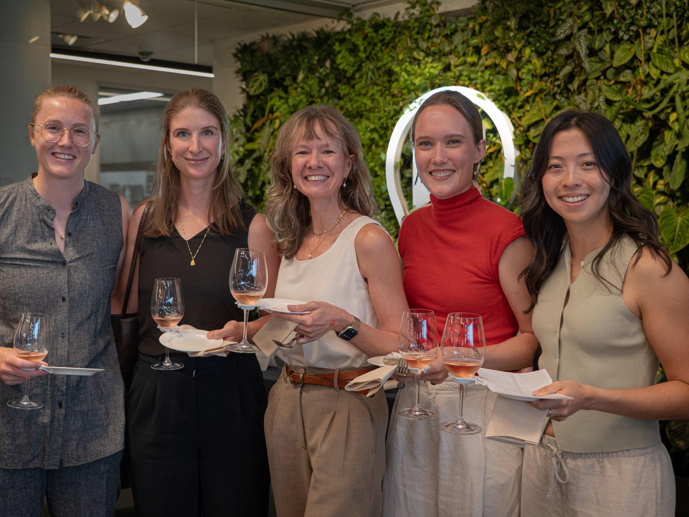 Five smiling women holding wine glasses and appetizers pose in front of a green wall with an illuminated circle.