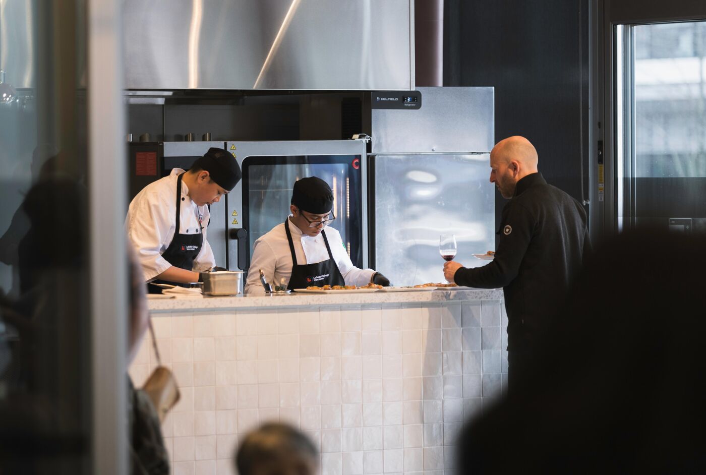 Two chefs are working behind a counter in a restaurant kitchen, while a customer drinks wine on the right.