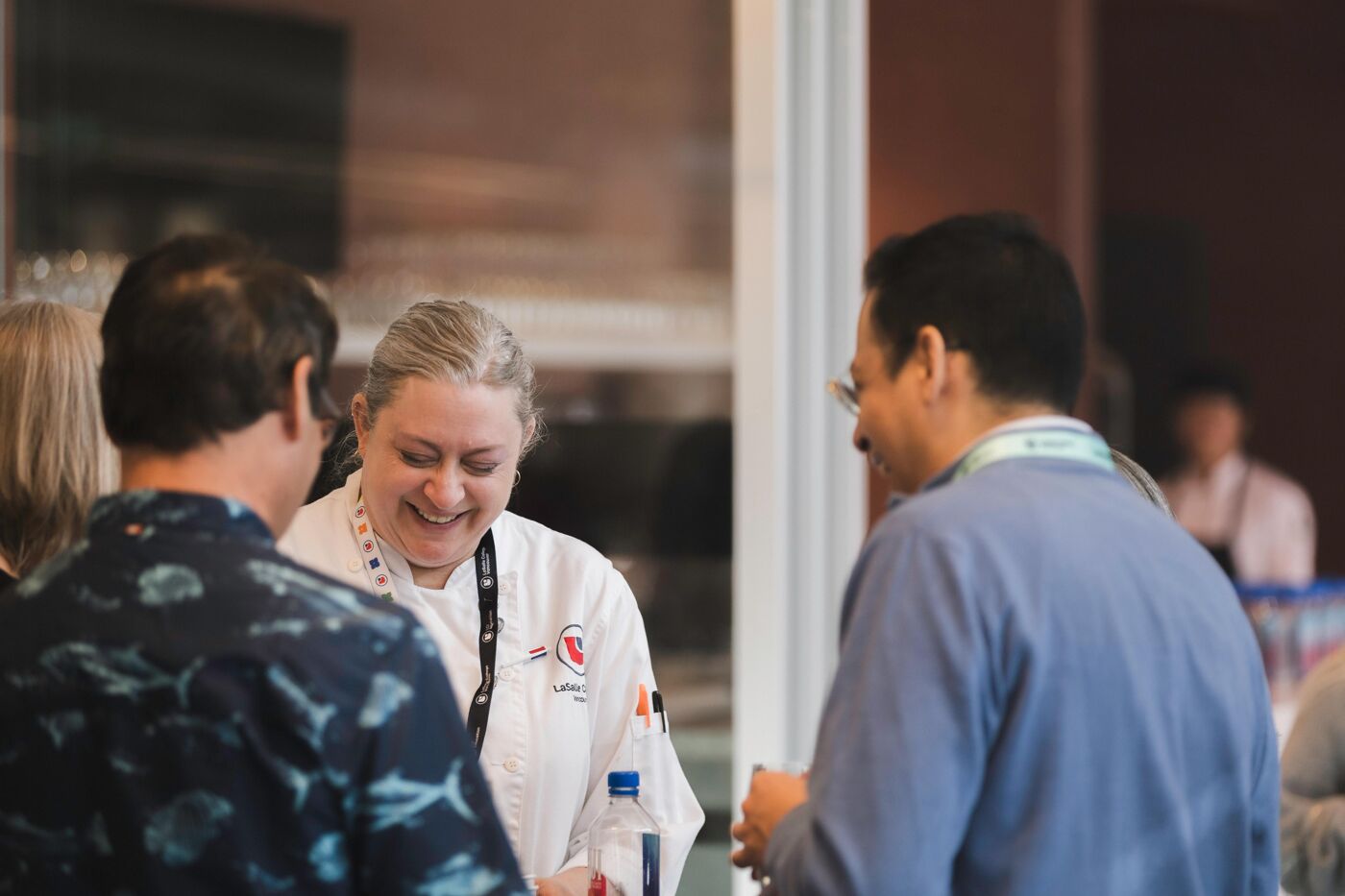 Three people are talking and smiling at an event. A woman in a chef's jacket appears to be explaining something to the two men in front of her.