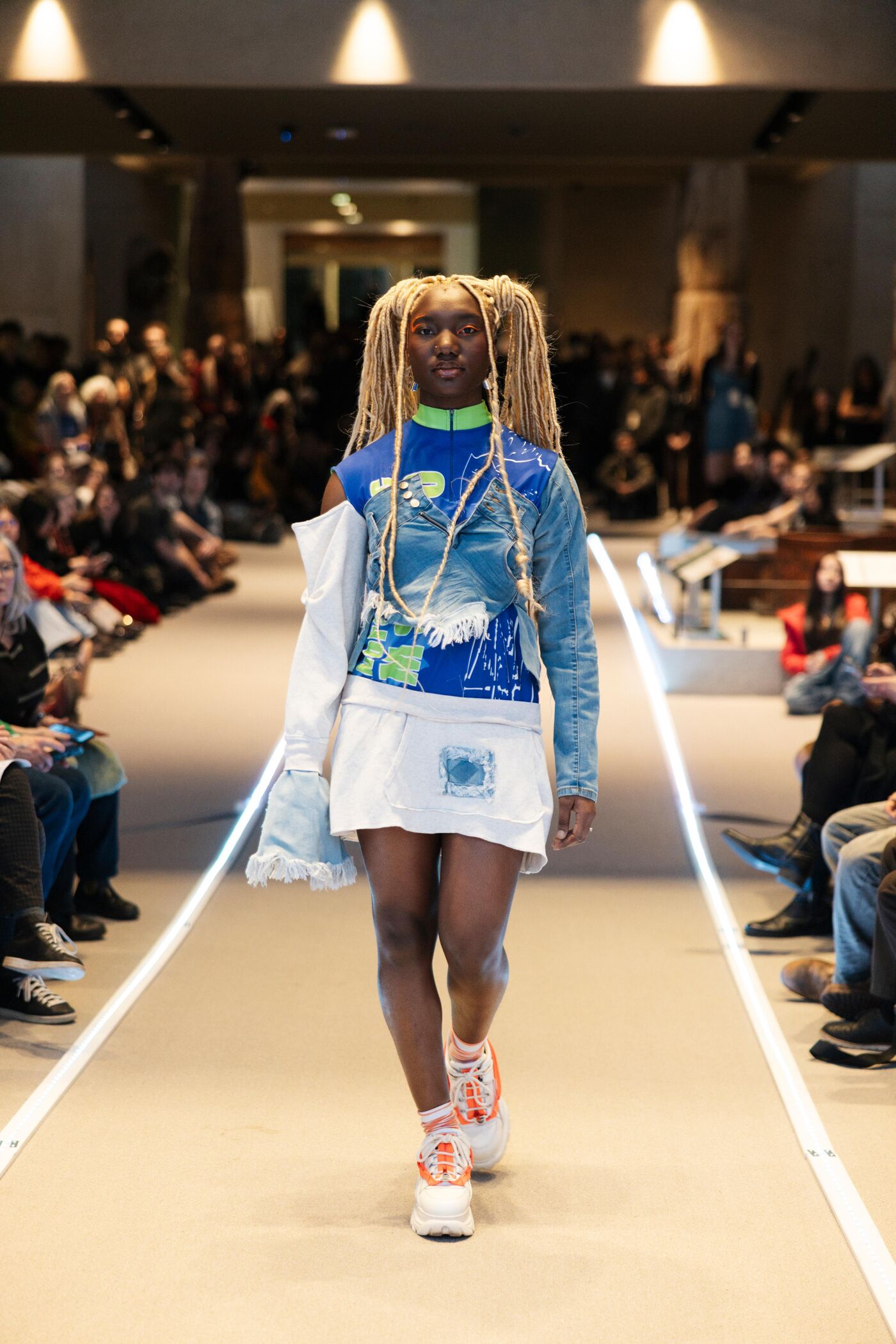 A woman with long dreadlocks and fashionable clothing walks down a runway in front of a seated audience. Her outfit includes a blue and green shirt, a white skirt, and sneakers.