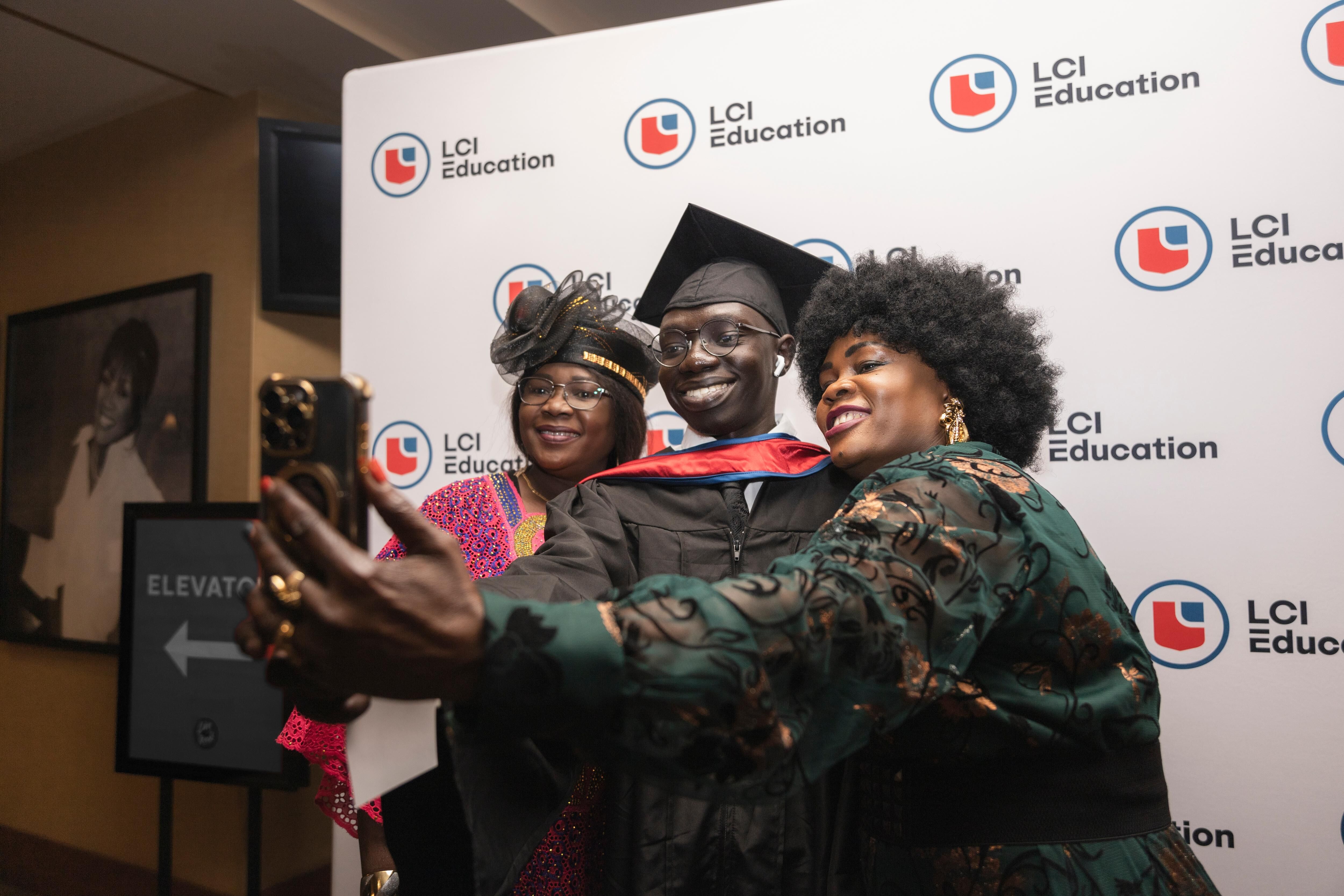 Three people are taking a selfie in front of a backdrop featuring the LCI Education logo. A man in graduation attire smiles as two women beside him also pose for the photo.