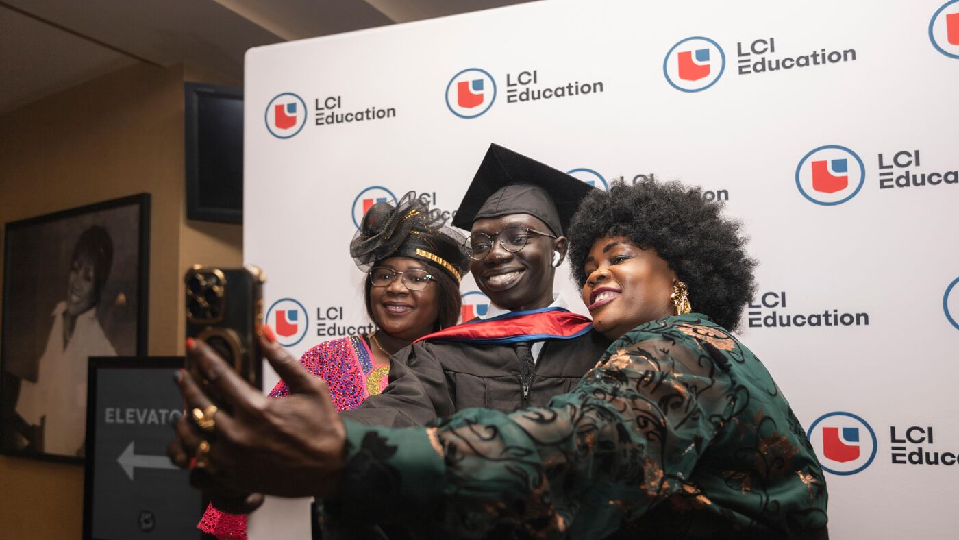 Three people are taking a selfie in front of a backdrop featuring the LCI Education logo. A man in graduation attire smiles as two women beside him also pose for the photo.