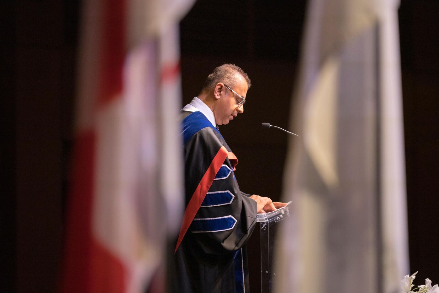 A man wearing an academic gown is reading a speech at a lectern during a ceremony. The man is wearing glasses and appears focused on his papers.