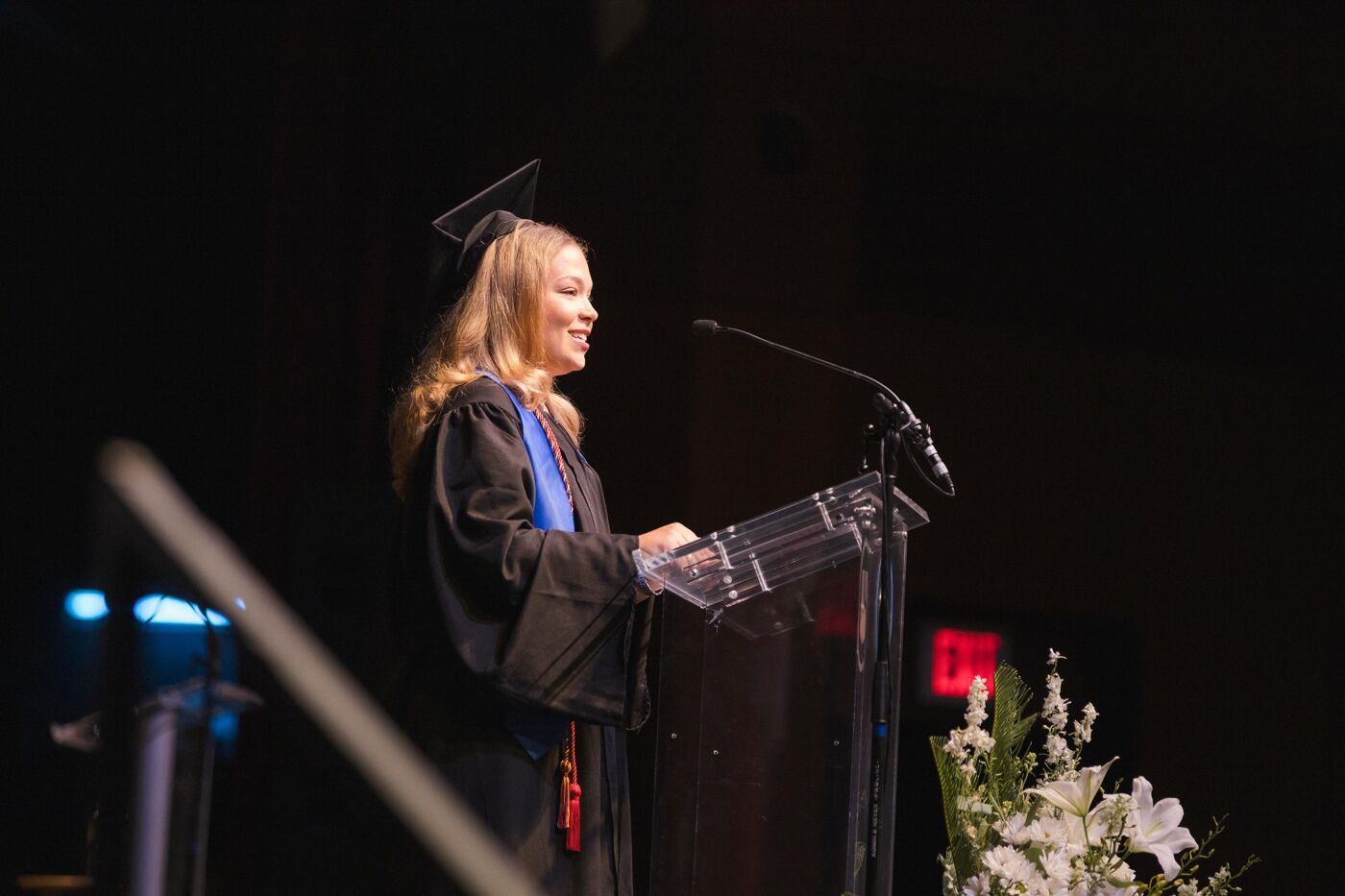 A smiling female graduate speaks at a podium during a commencement ceremony, dressed in a cap and gown.