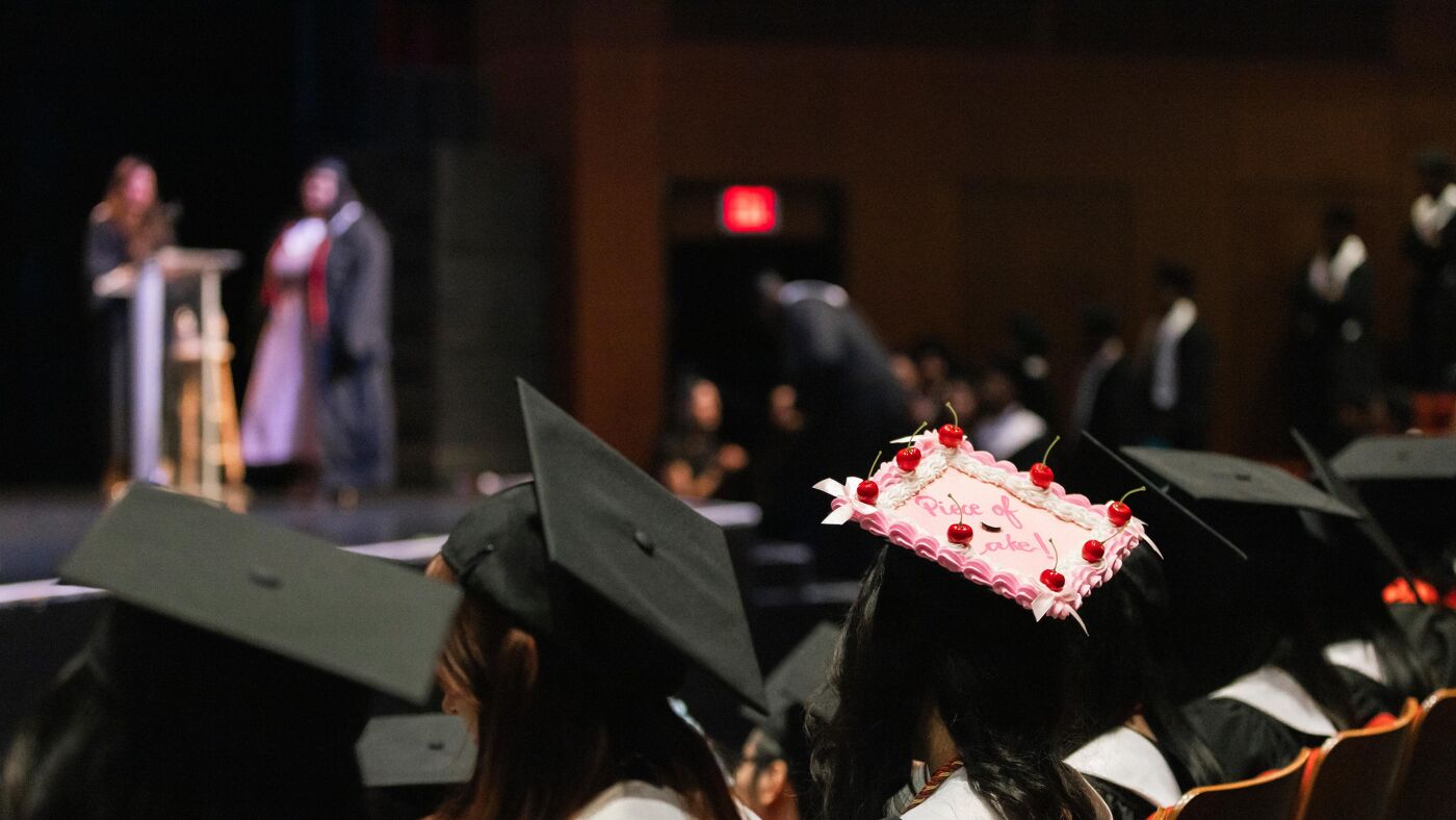 A view from behind of a group of graduates wearing caps and gowns, seated in rows, facing a stage where blurred figures are visible.