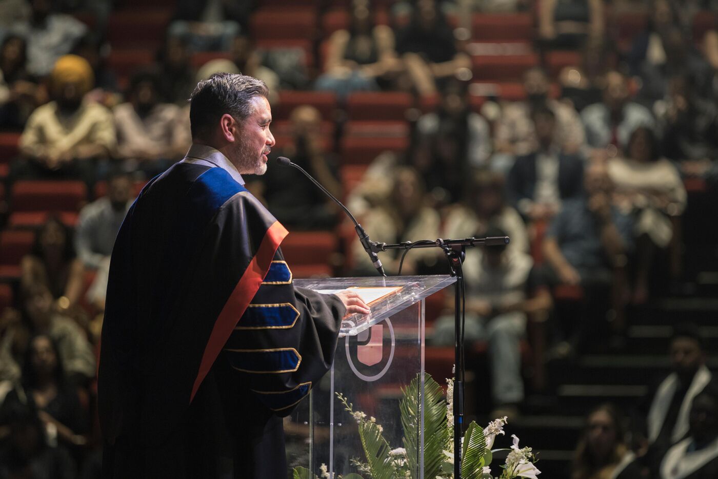 A man in academic regalia speaks from a podium in front of an audience in an auditorium, possibly during a graduation ceremony or academic address.