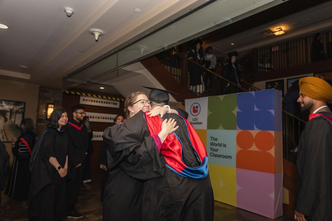 Two graduates embrace joyfully, dressed in academic gowns and caps, celebrating their achievement on a special day. Other students and guests are also visible in the background.
