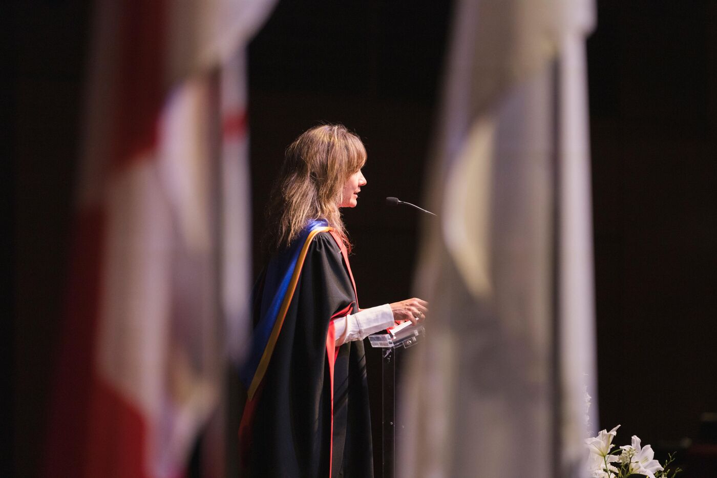 A woman in academic regalia speaks at a microphone, possibly during a graduation ceremony, with a dark background and columns in the foreground.
