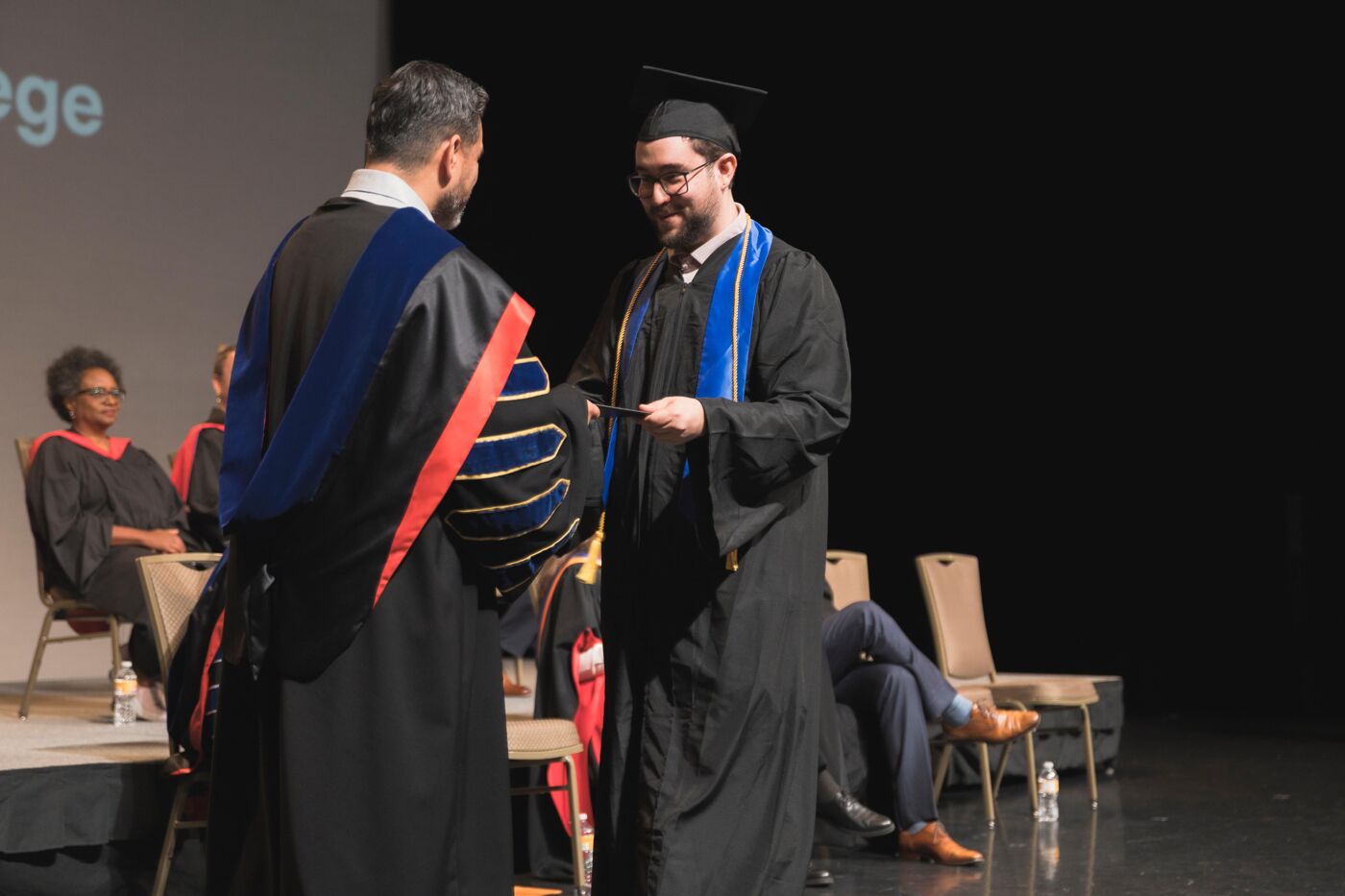 A student receives their diploma during a graduation ceremony, dressed in a cap and gown, as a university official hands them the document.