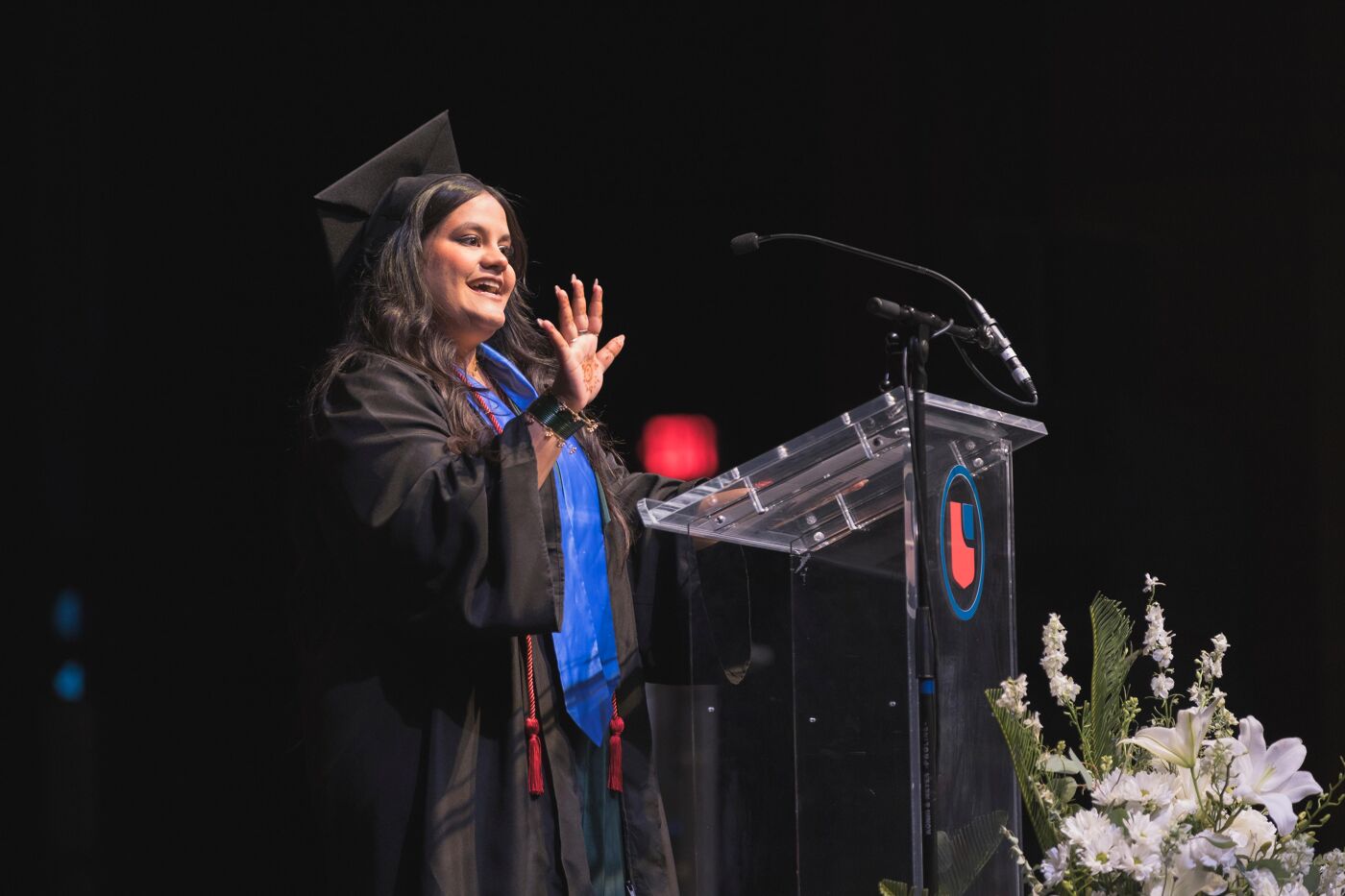 A female graduate speaks at a podium during a commencement ceremony, wearing a cap and gown, with a microphone in front of her.