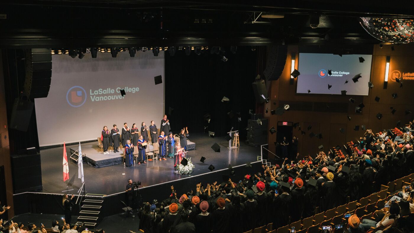 A graduation ceremony at LaSalle College Vancouver, with students tossing their caps in the air as an audience applauds.