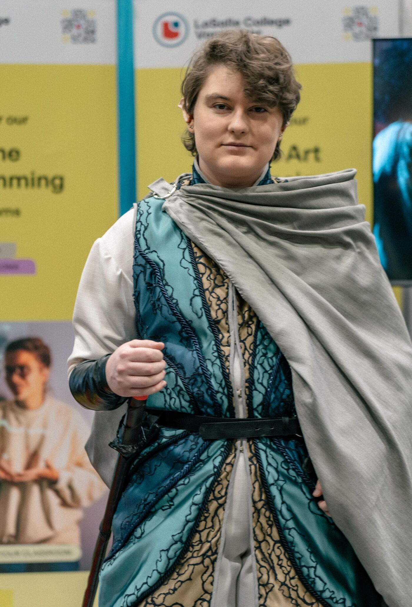 A person in an elaborate costume, possibly fantasy or movie-themed, stands against a yellow backdrop with text. The costume features intricate details and a cape.
