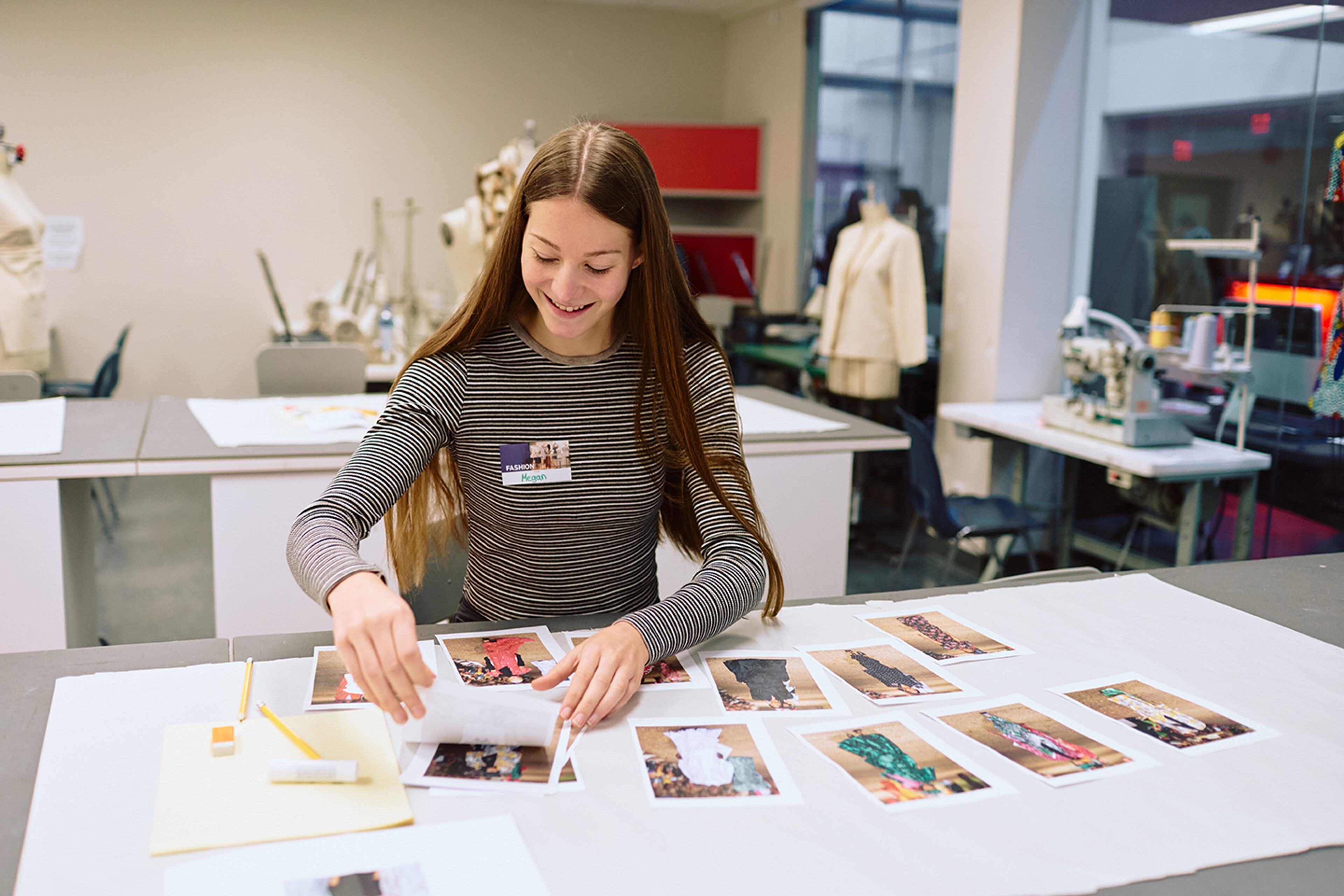 A smiling fashion design student organizes her garment sketches and fabric samples on a worktable in a bright studio.