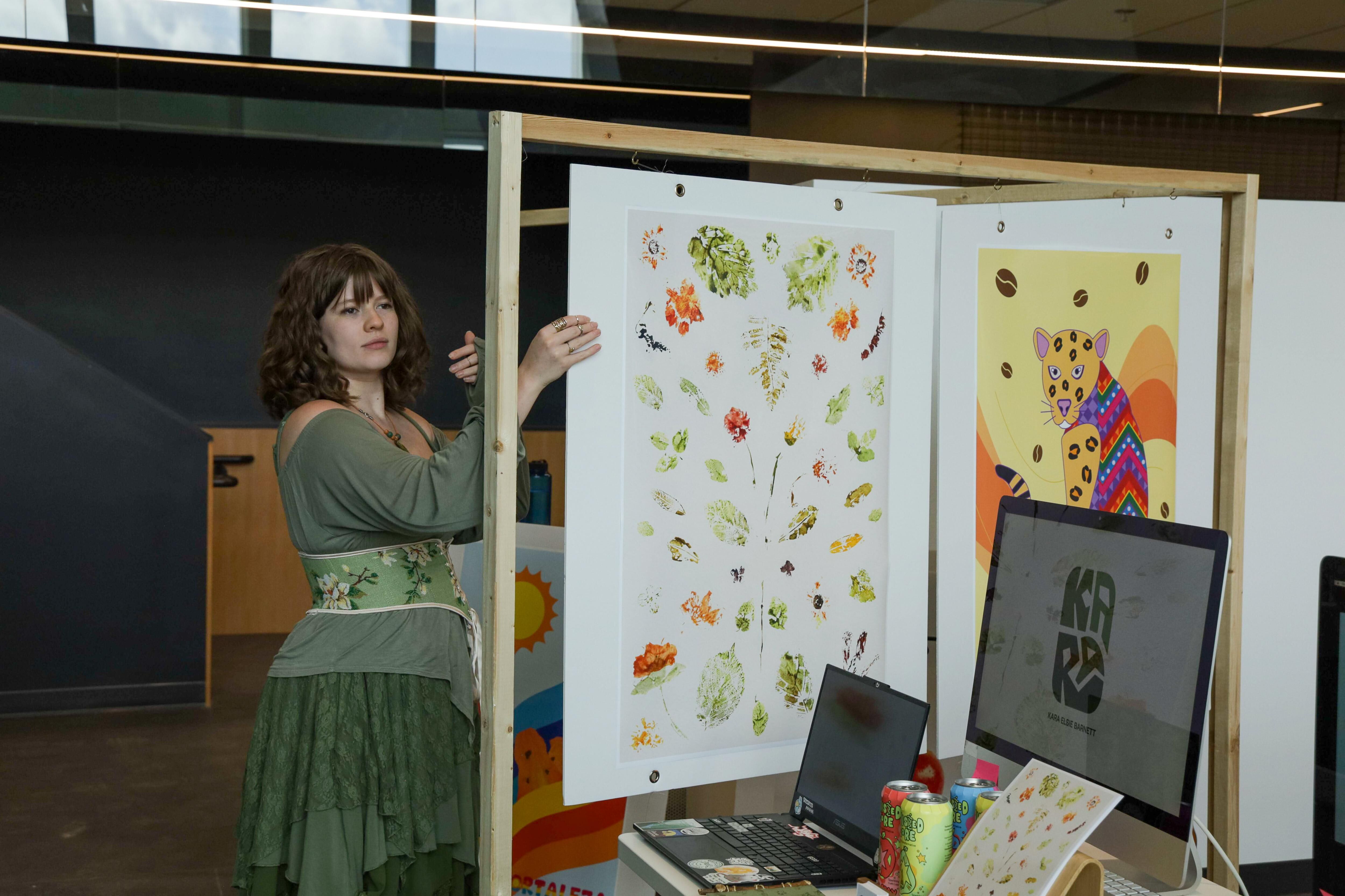 A woman in a green dress stands next to a display of artwork. The artwork includes botanical prints and a colorful abstract image of a leopard. A laptop and monitor are also visible on the table.