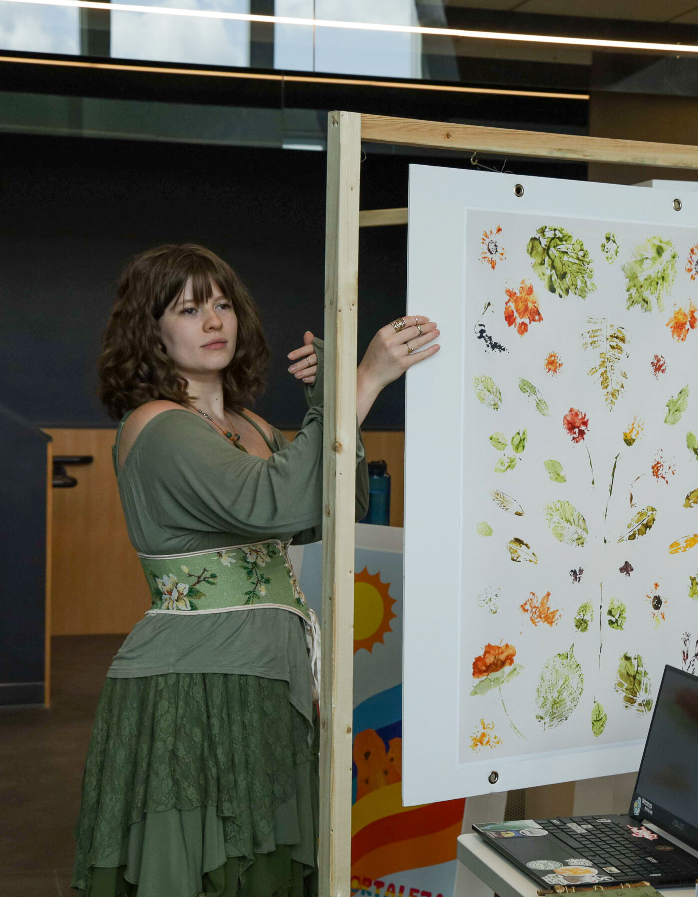 A woman in a green dress stands next to a display of artwork. The artwork includes botanical prints and a colorful abstract image of a leopard. A laptop and monitor are also visible on the table.