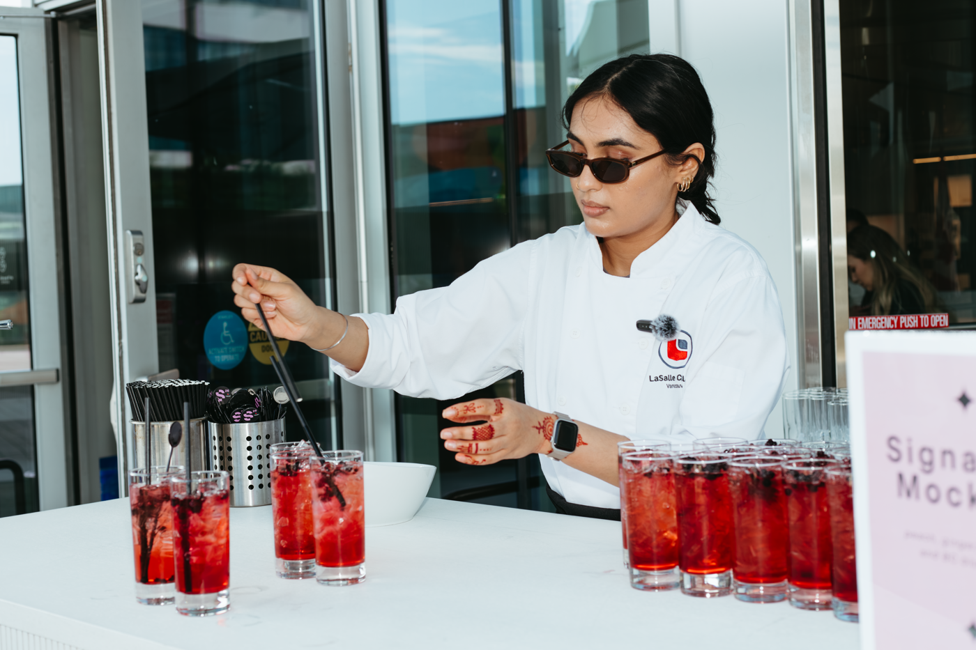 A woman wearing sunglasses and a chef's jacket prepares red drinks with fruit and straws at an event.