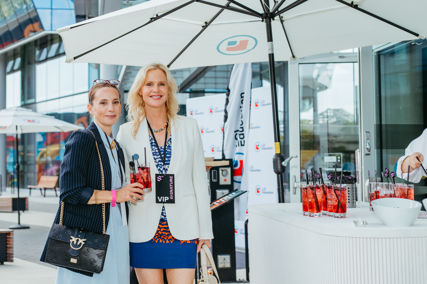 Two women holding red drinks stand under a white umbrella in front of a bar with more beverages. The woman on the right wears a white jacket and a VIP pass.