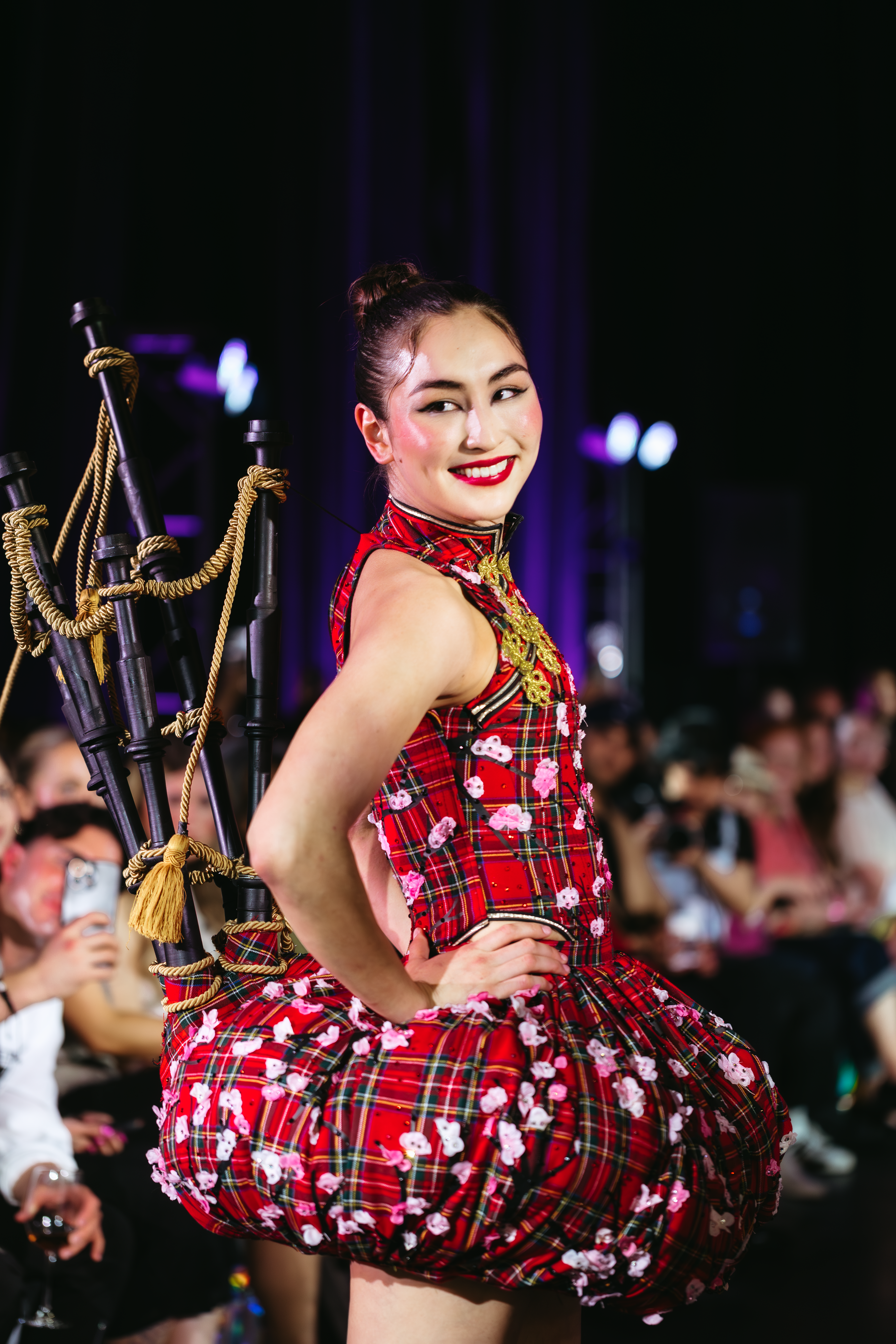 A smiling woman in a red tartan dress adorned with white flowers, featuring a bagpipe-like accessory on her back, poses on a runway.