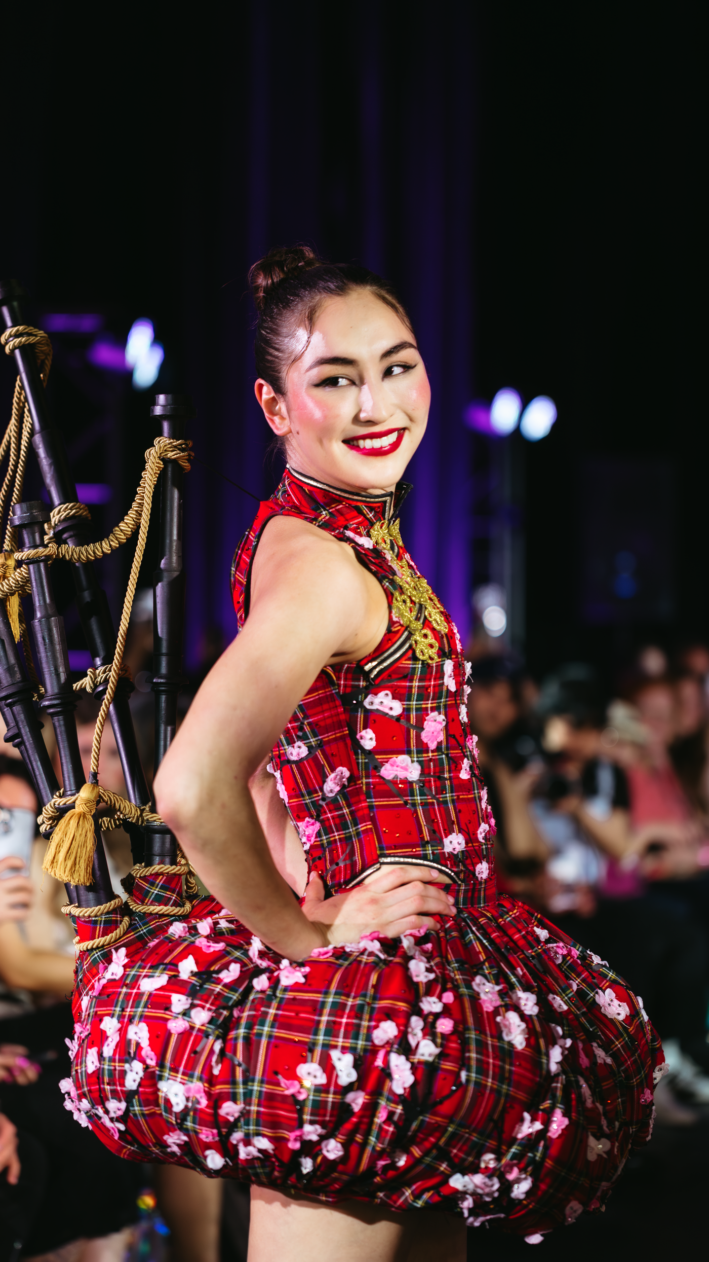 A smiling woman in a red tartan dress adorned with white flowers, featuring a bagpipe-like accessory on her back, poses on a runway.