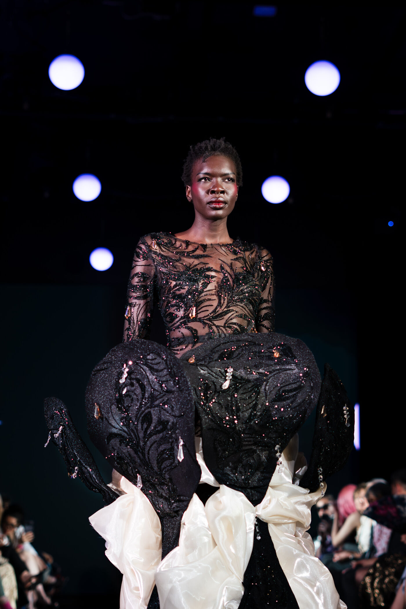 A model showcases a black dress with feather-like details and a voluminous skirt design, possibly during a fashion show.