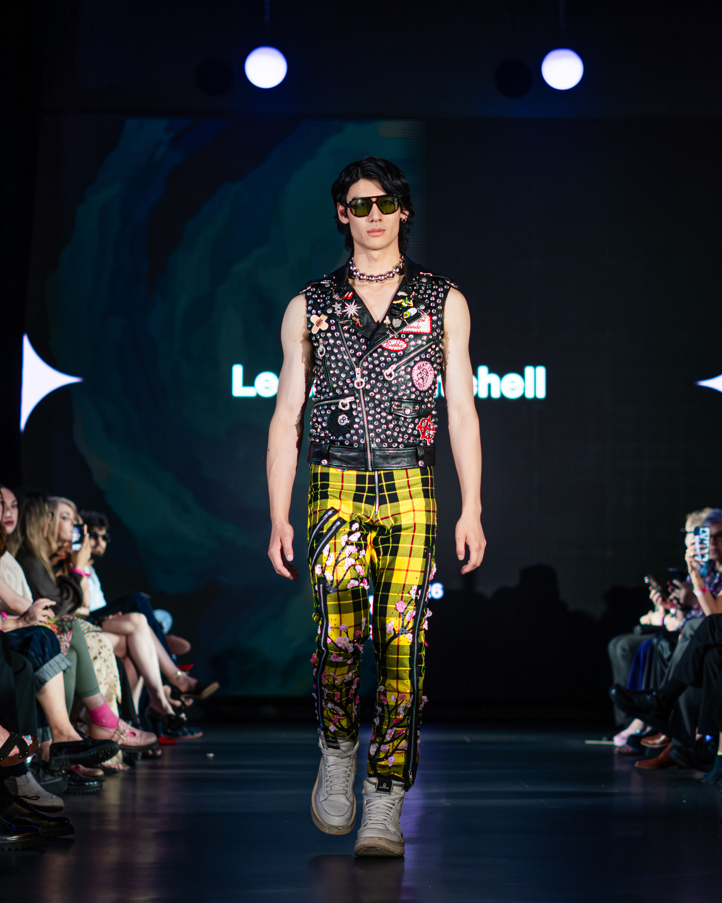 A male model walks down the runway in a punk rock outfit featuring a vest, yellow plaid pants, and silver accessories.