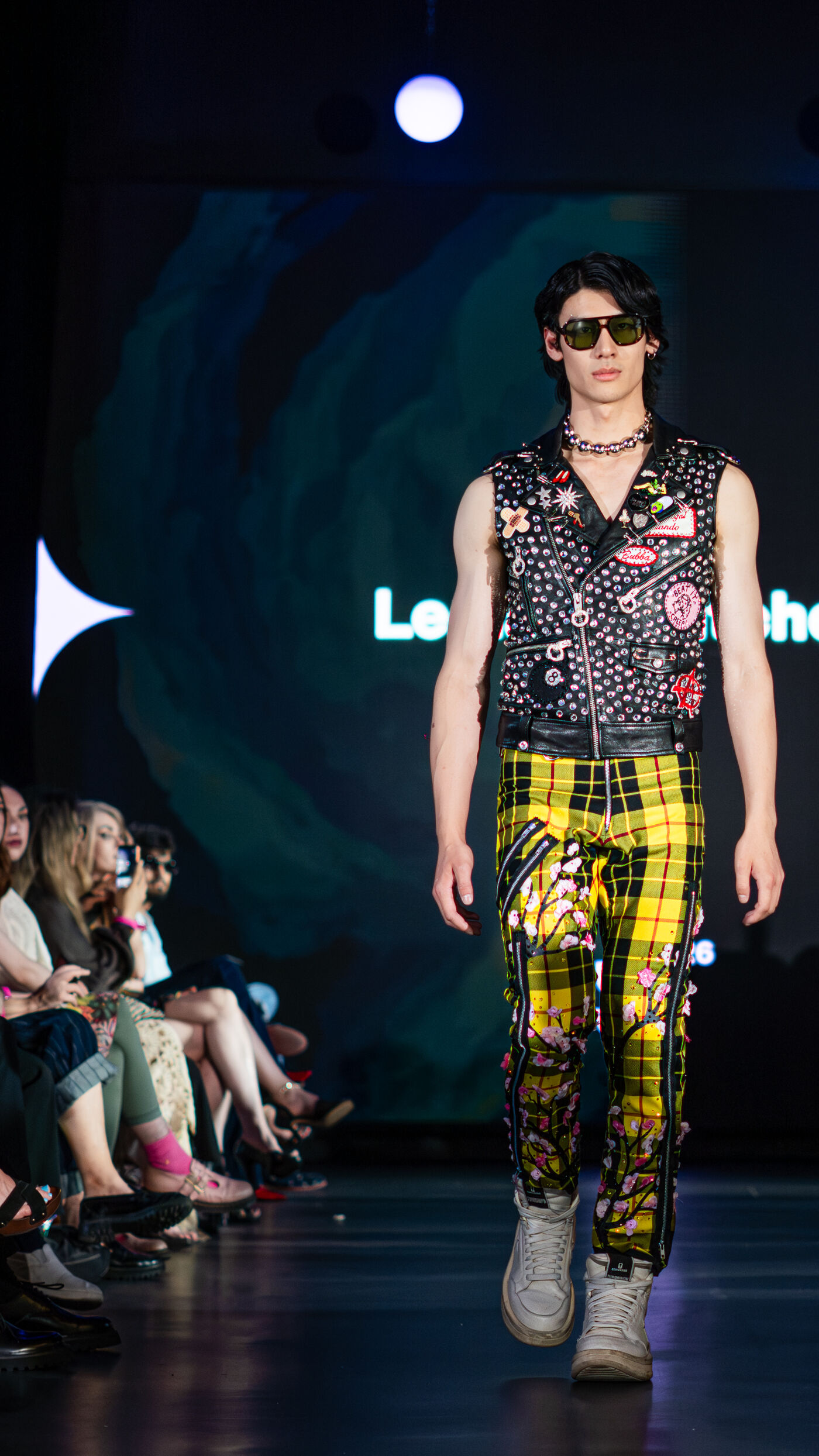 A male model walks down the runway in a punk rock outfit featuring a vest, yellow plaid pants, and silver accessories.