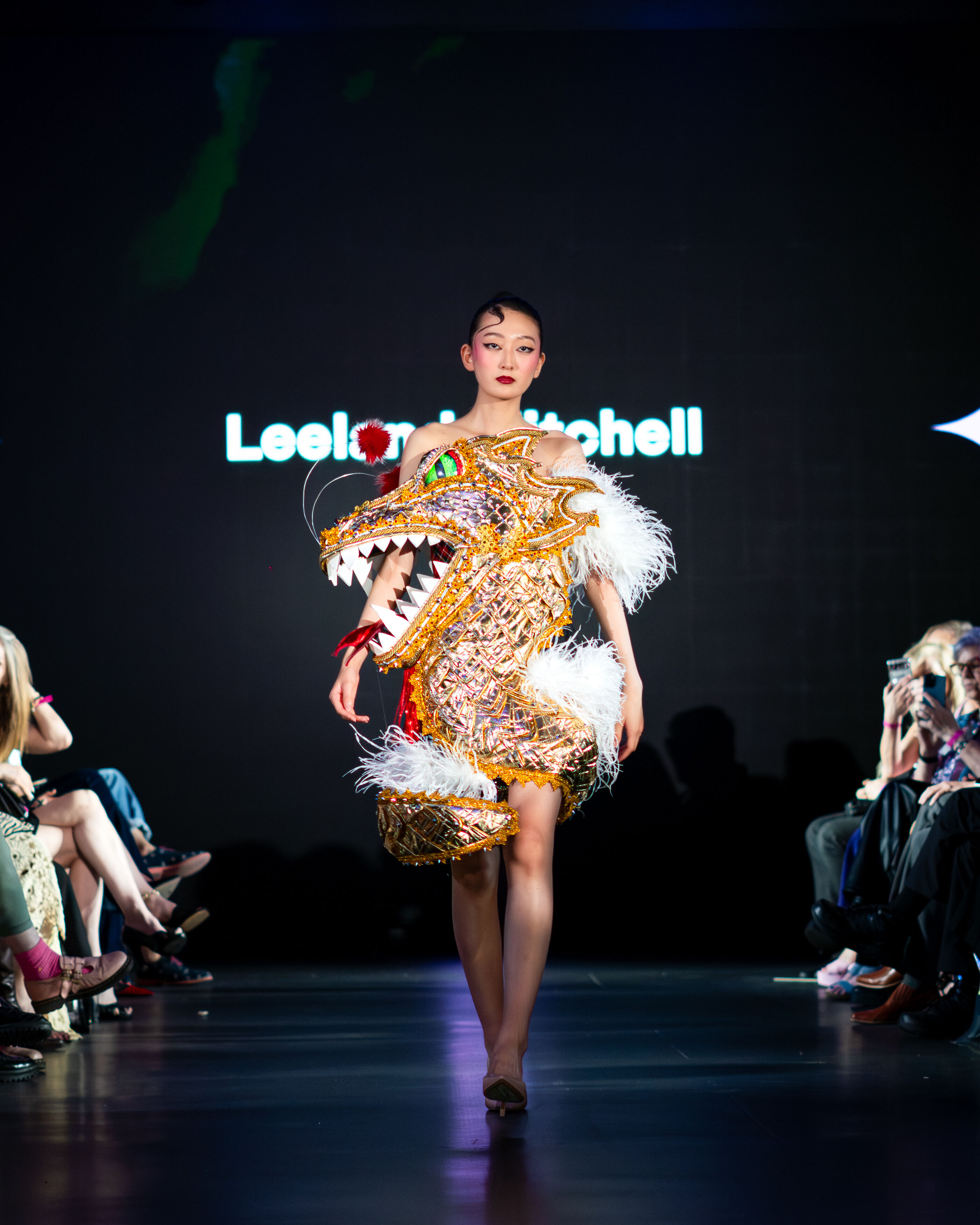 A model walks the runway in a spectacular golden dragon-shaped dress, featuring white feathers and striking details.