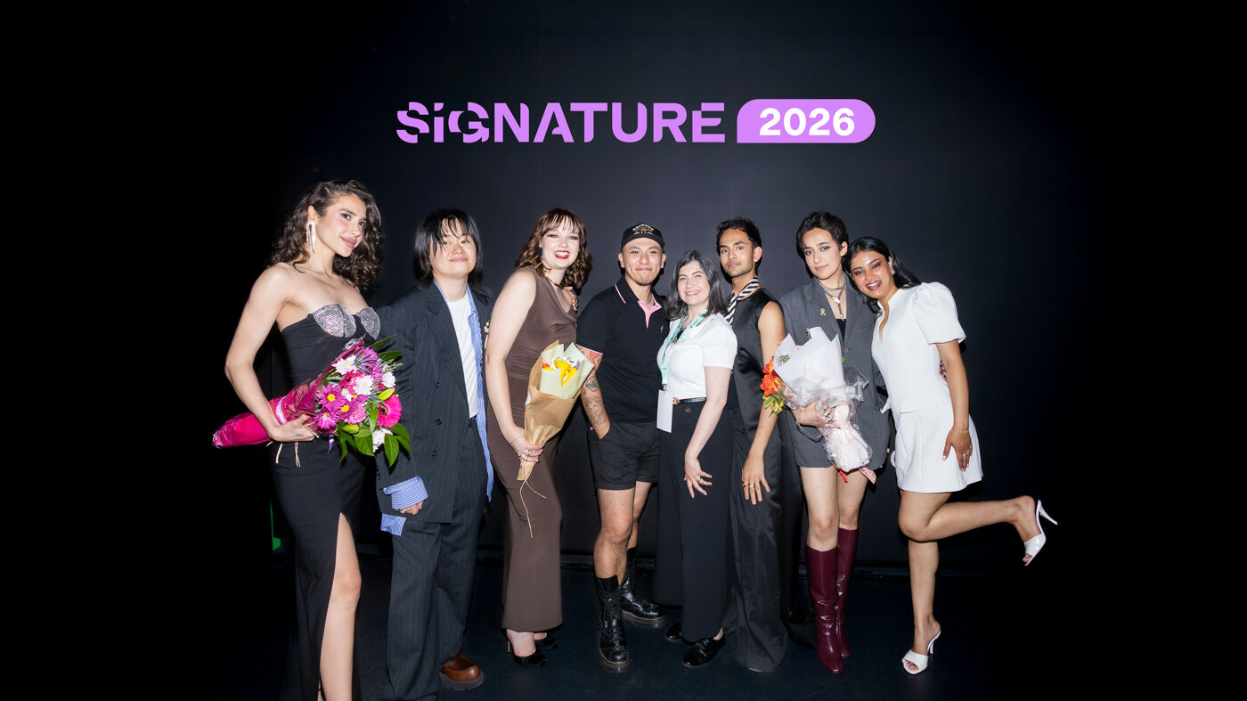 A group of nine people pose for a photo in front of a black backdrop featuring the "SIGNATURE 2026" logo at the top. Several individuals are holding bouquets of flowers.