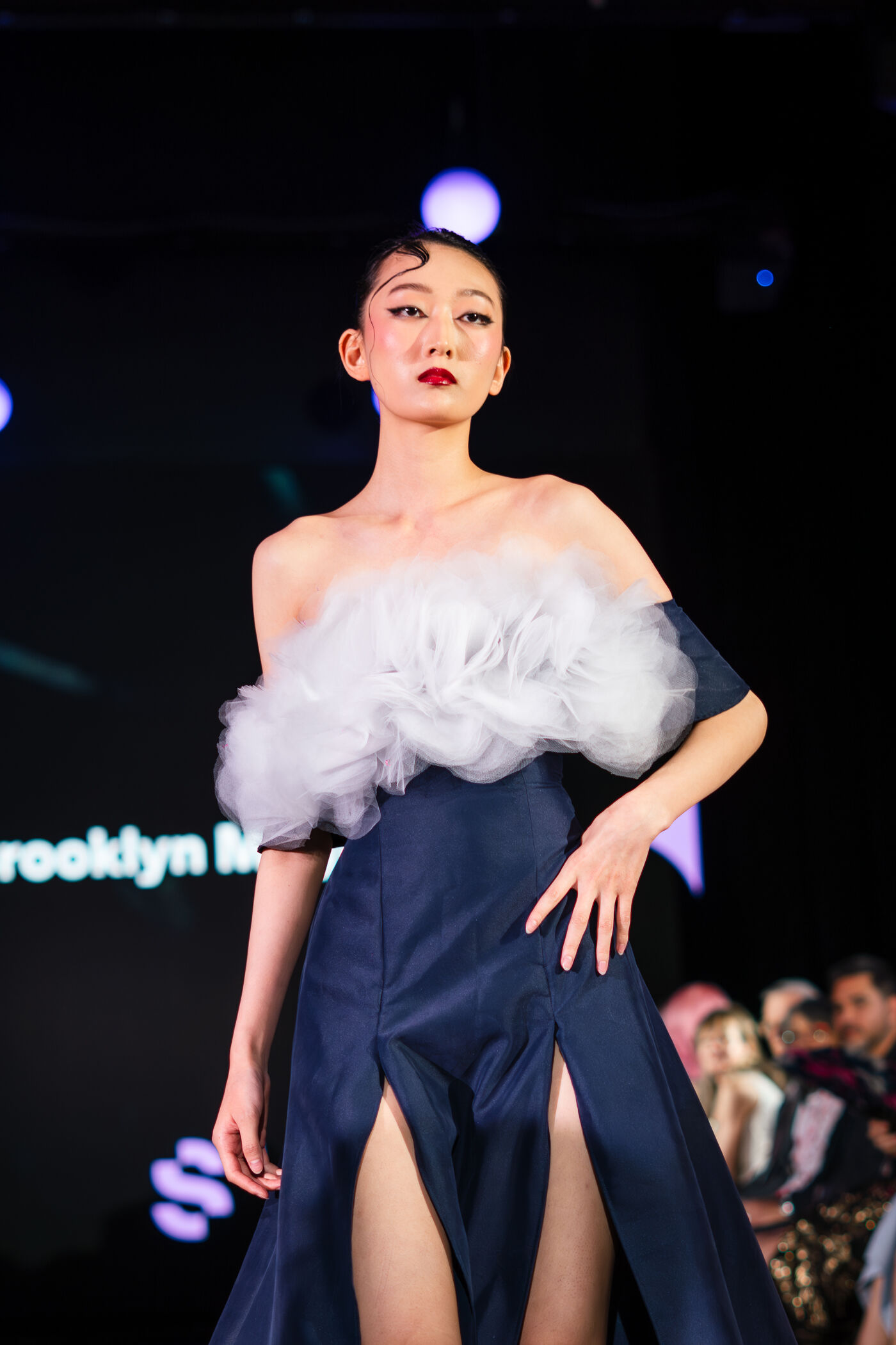A young woman in a dark blue dress with thigh-high slits and a voluminous white detail on top, posing on a runway.