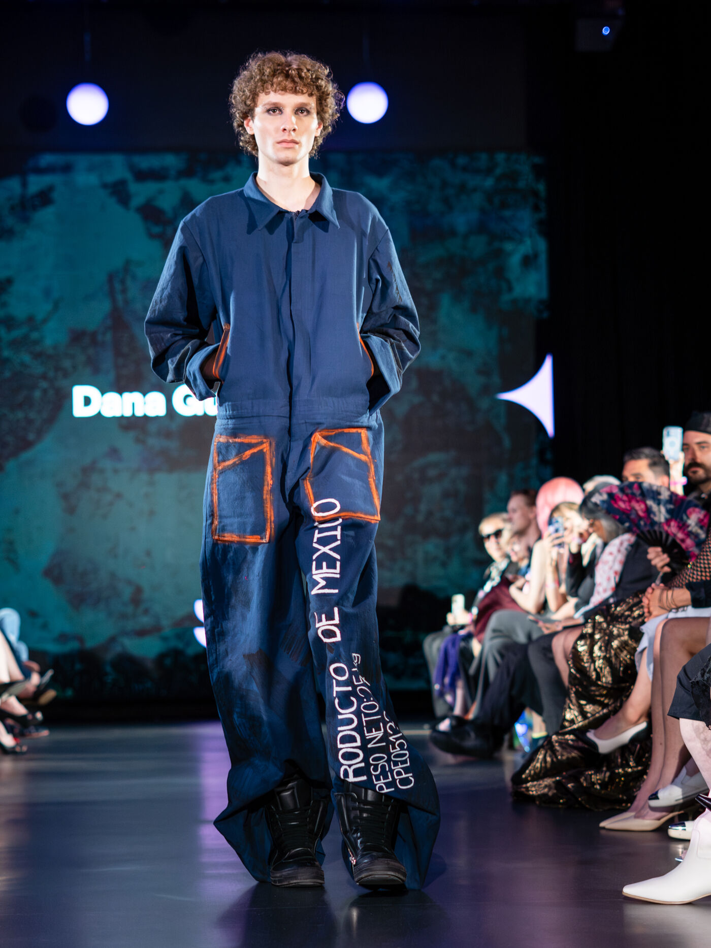 A male model walks the runway in a blue jumpsuit featuring orange pockets and printed text, with an audience seated on either side.