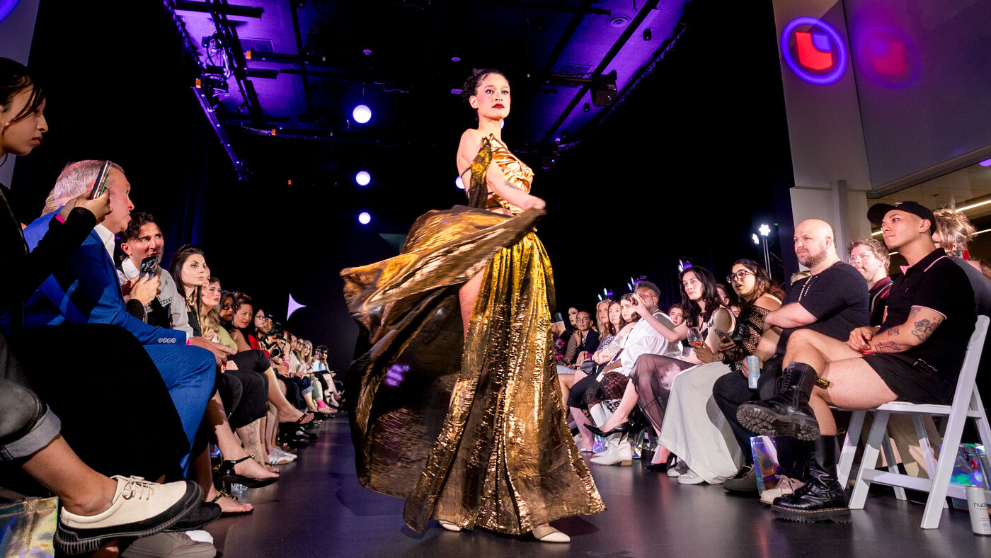 A female model showcases a long, shimmering gold gown with a thigh-high slit, walking down a runway in front of a seated audience.