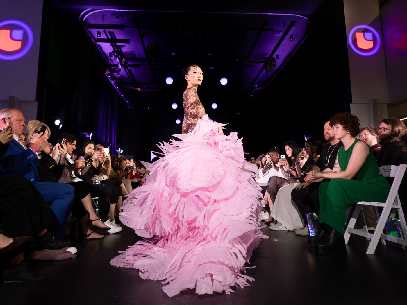 A model walks down a runway in a dramatic pink ruffled gown, with an audience applauding in the background under purple lighting.