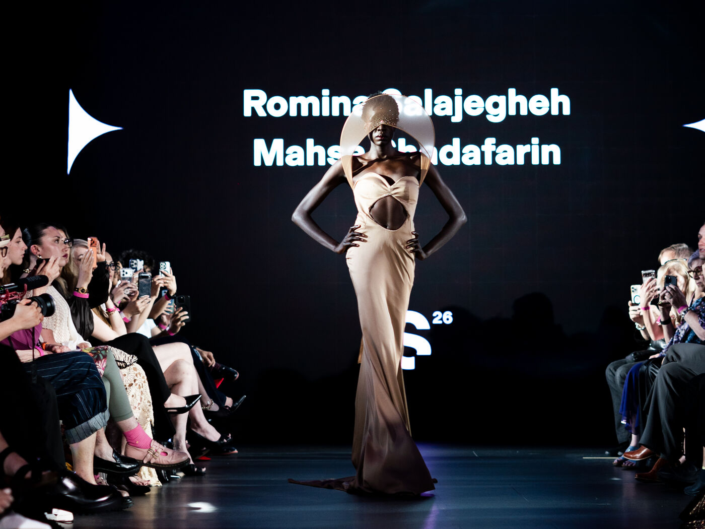 A model showcases a long, form-fitting gown with an oversized hat on a runway, while an audience applauds on either side.
